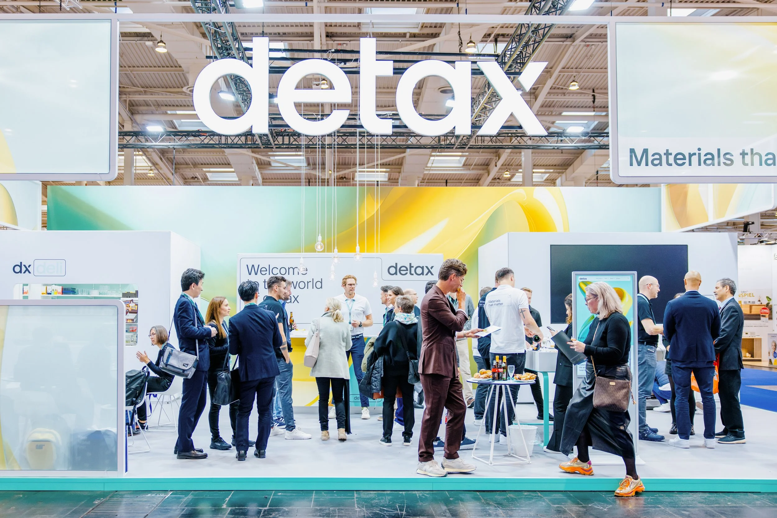 Messestand mit dem großen Schriftzug 'detax', auf dem viele Menschen in Business-Kleidung stehen und Gespräche führen, einige schauen auf Tablets oder Broschüren, im Hintergrund leuchtende Kabel und moderne Präsentationselemente.