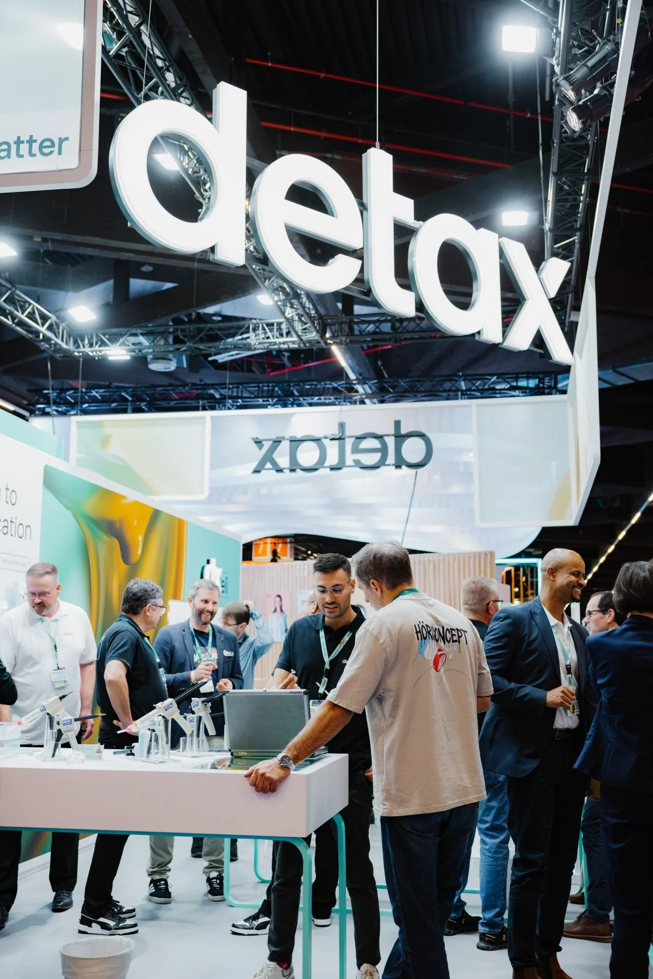 Auf einer Messe mit einer großen leuchtenden Beschilderung, die das Wort 'detax' zeigt, sind mehrere Menschen im Gespräch und betrachten Produkte auf einem Stand. Im Vordergrund steht ein Mann in einem beige T-Shirt mit der Aufschrift 'HÖRCONCEPT', der mit zwei anderen Personen diskutiert. Im Hintergrund sind weitere Besucher, die sich unterhalten.
