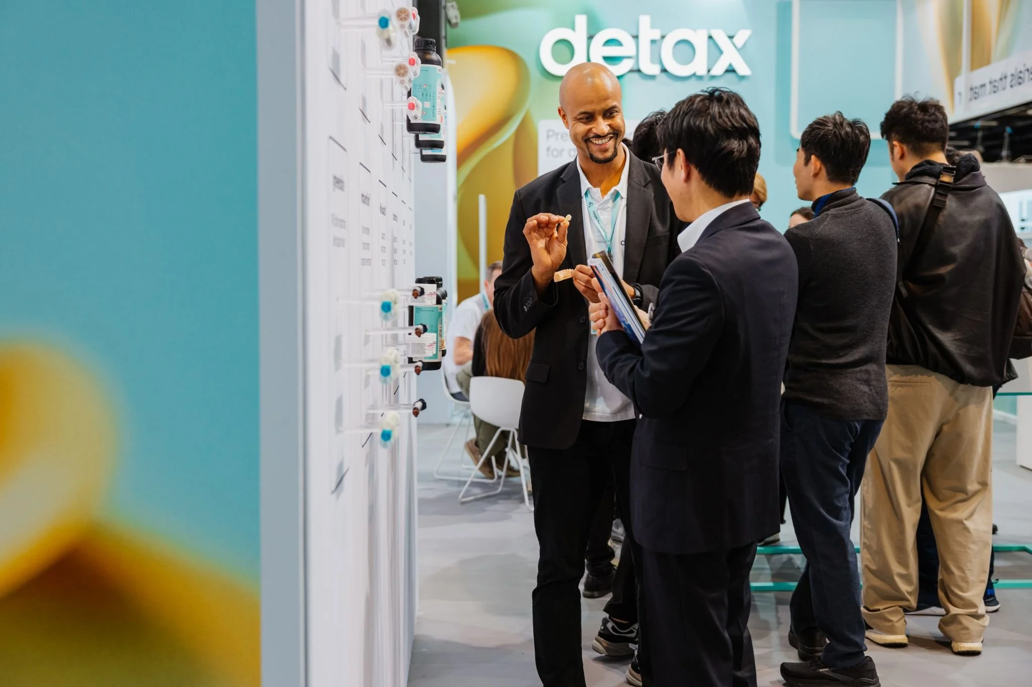 Mehrere Menschen unterhalten sich in einer Ausstellungshalle. Ein Mann zeigt auf etwas, während ein anderer mit einem Tablet spricht. Im Hintergrund ein großes Schild mit dem Wort "detax".