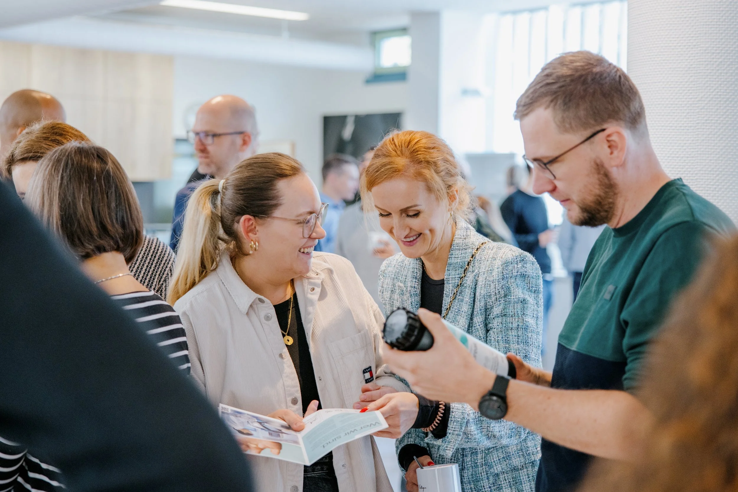 Menschen bei einer Veranstaltung, die Spaß haben und sich unterhalten, einige schauen auf Broschüren. Im Hintergrund weitere Personen in einem Raum mit natürlichem Licht.
