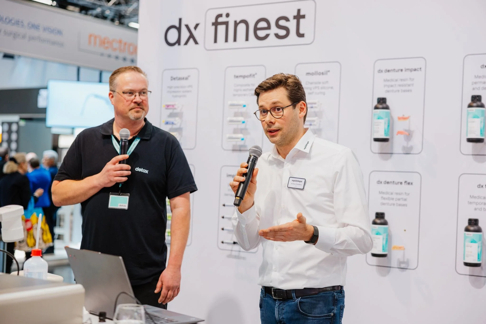 Zwei Männer sprechen auf einer Messe vor einem Display mit Produkten der Marke dx finest. Einer trägt ein schwarzes T-Shirt mit dem Logo detax, der andere eine weiße Hemd mit Namensschild und hält ein Mikrofon, während er spricht.