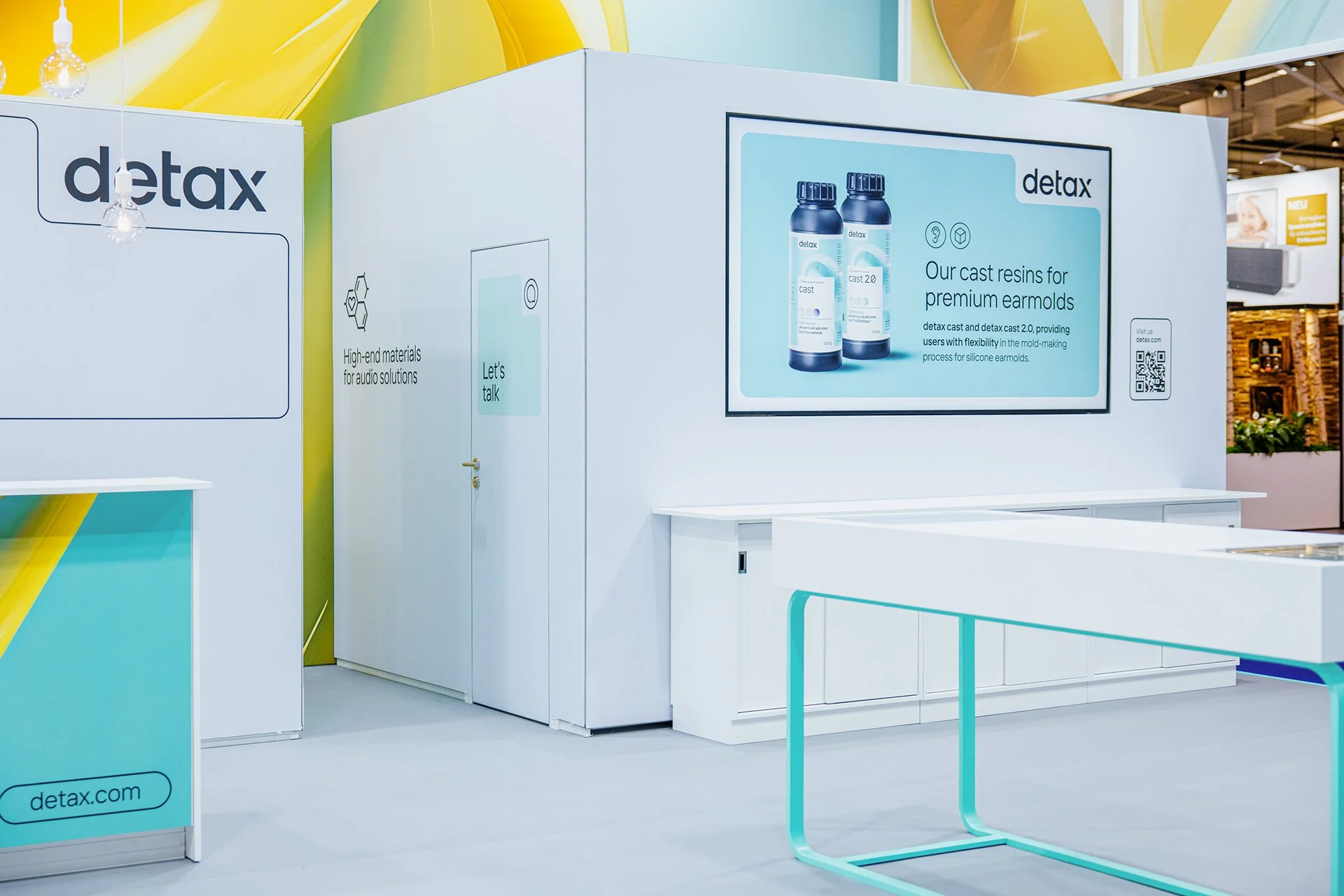 Messestand der Marke detax mit einem großen Bildschirm, der zwei Flaschen mit Resin für Ohrhörer zeigt und Informationen darüber gibt.