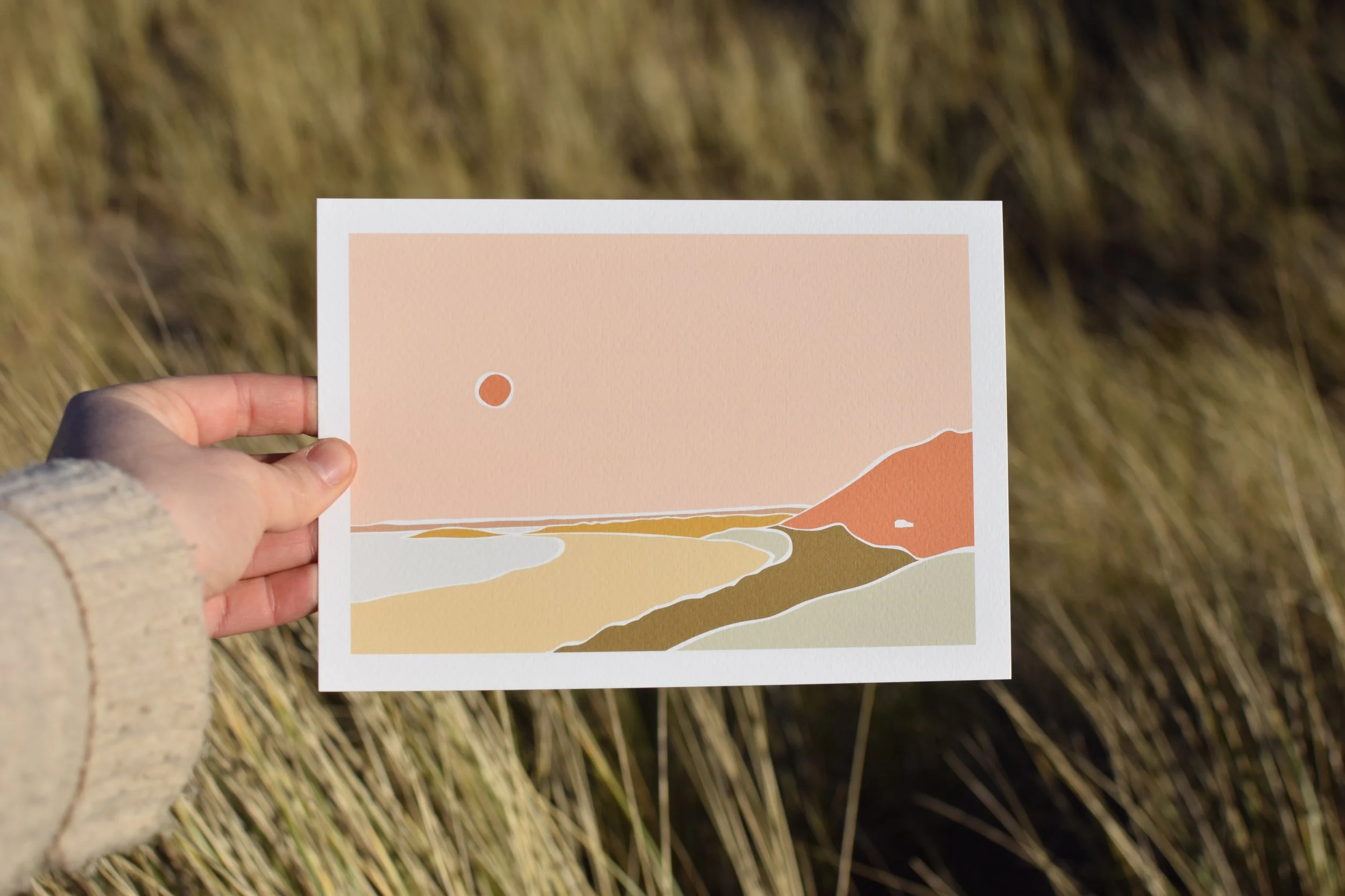 A person holds a pastel-colored illustrated landscape postcard featuring a sun, a hill, and a winding path against a grassy field background.