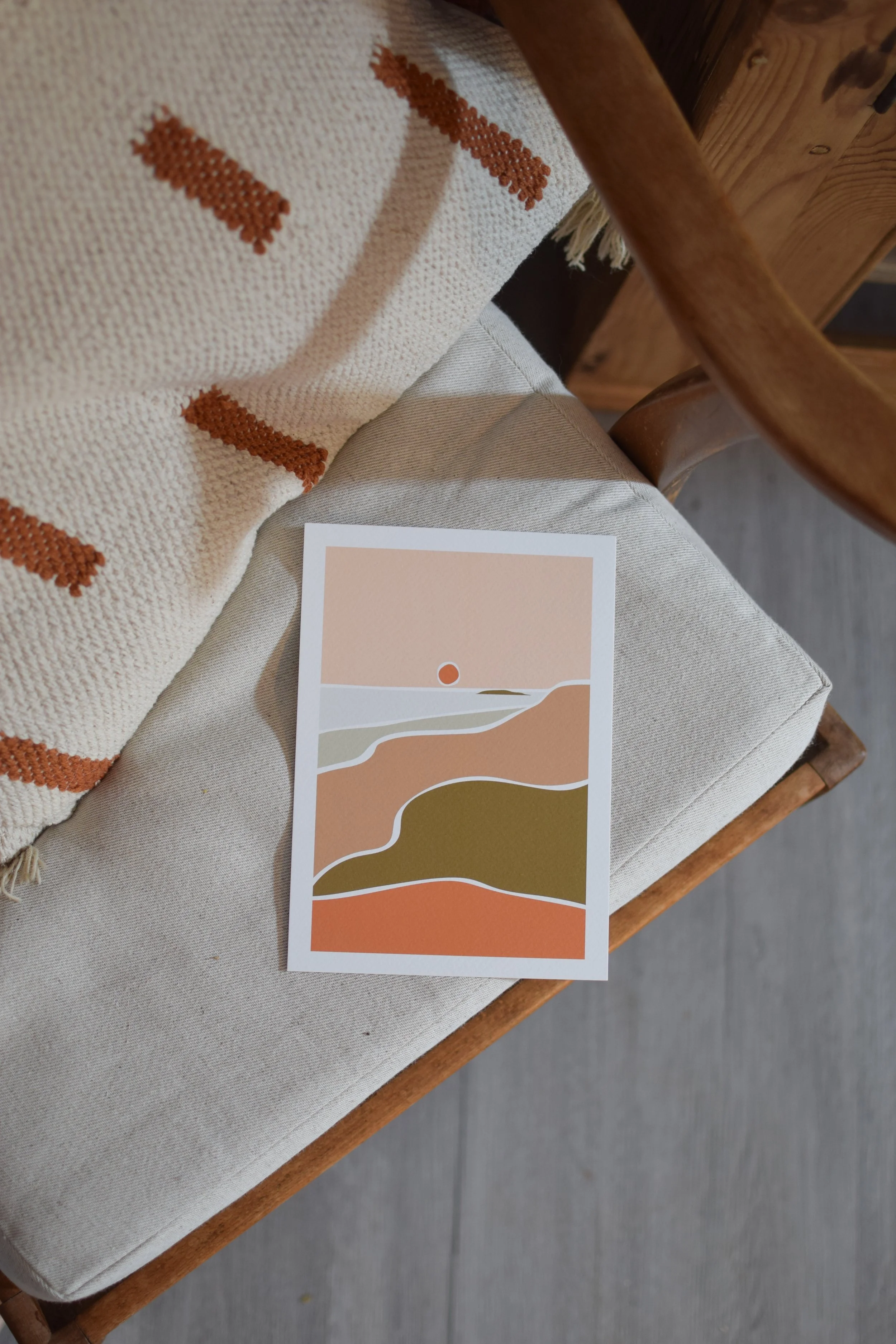 A minimalistic landscape illustration print on paper, featuring layers of land, water, and sky in earth tones, with a small red sun or moon above the horizon.