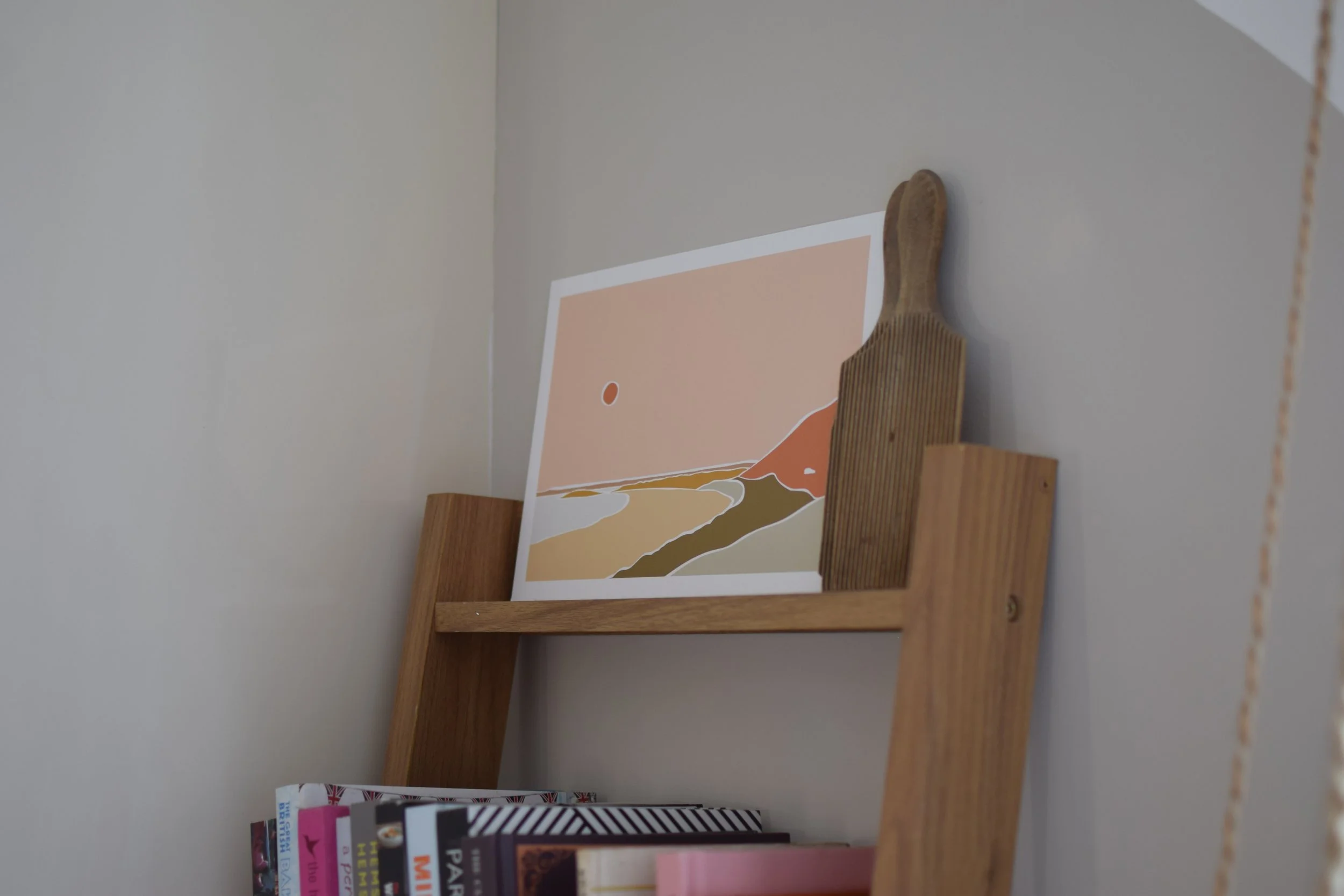 A wooden shelf holding a framed minimalist landscape art print with a house, a river, and a mountain, positioned above a row of books on a white wall.