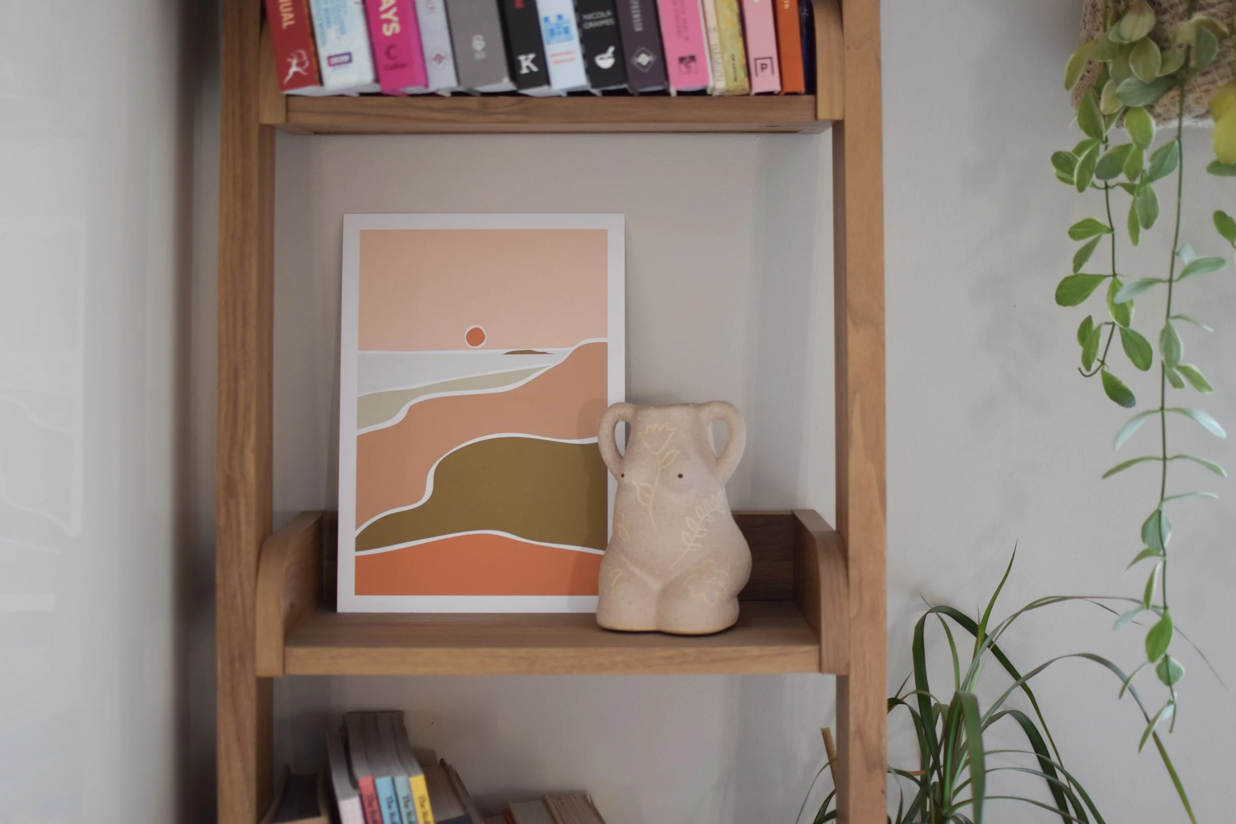 Decorative scene on a wooden bookshelf with a framed landscape art print, a beige decorative vase, a small stack of books on the lower shelf, and a trailing green houseplant.