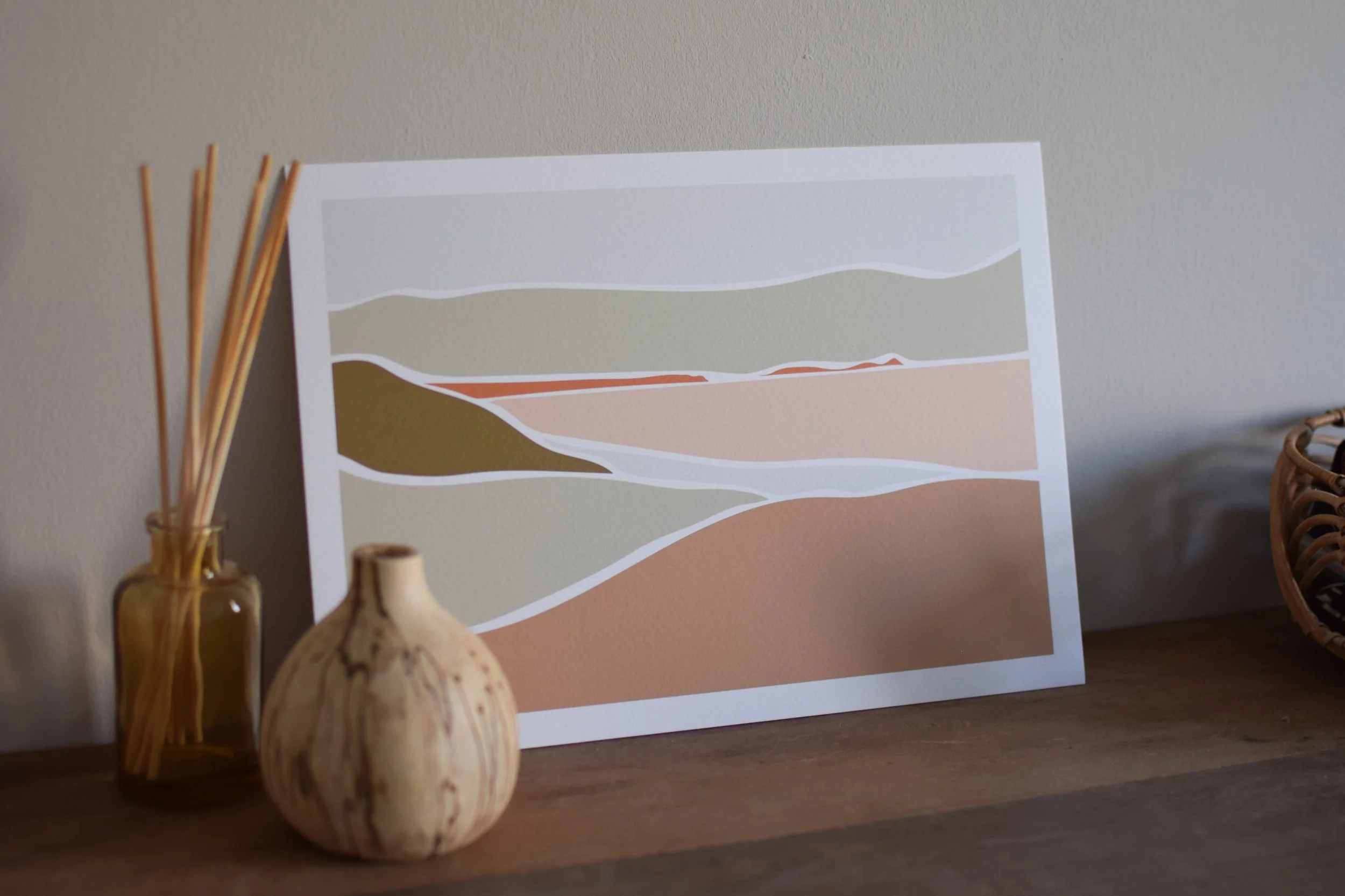 Decorative framed landscape art with abstract mountain and ground shapes in neutral tones, positioned on a wooden surface. To the left, a small yellow glass jar containing reed diffuser sticks, and in front, a rounded stone vase or sculpture with a n