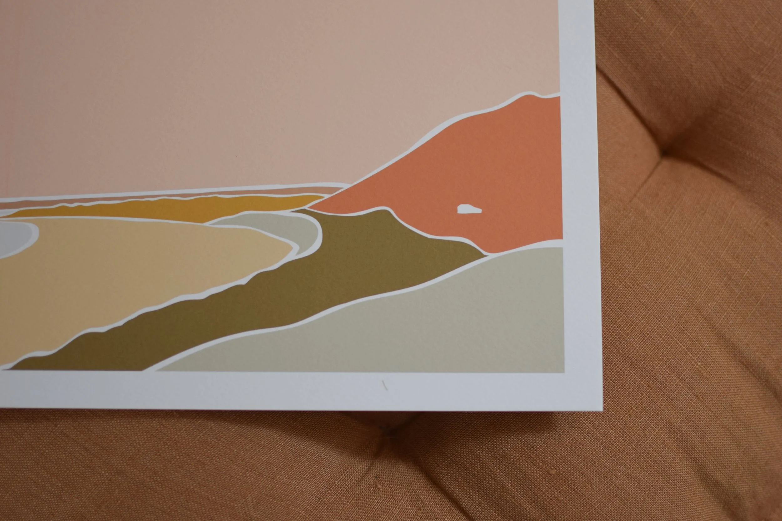 A stylized landscape illustration with layered hills and mountains in earth tones on a pastel pink background, resting on a brown fabric surface.