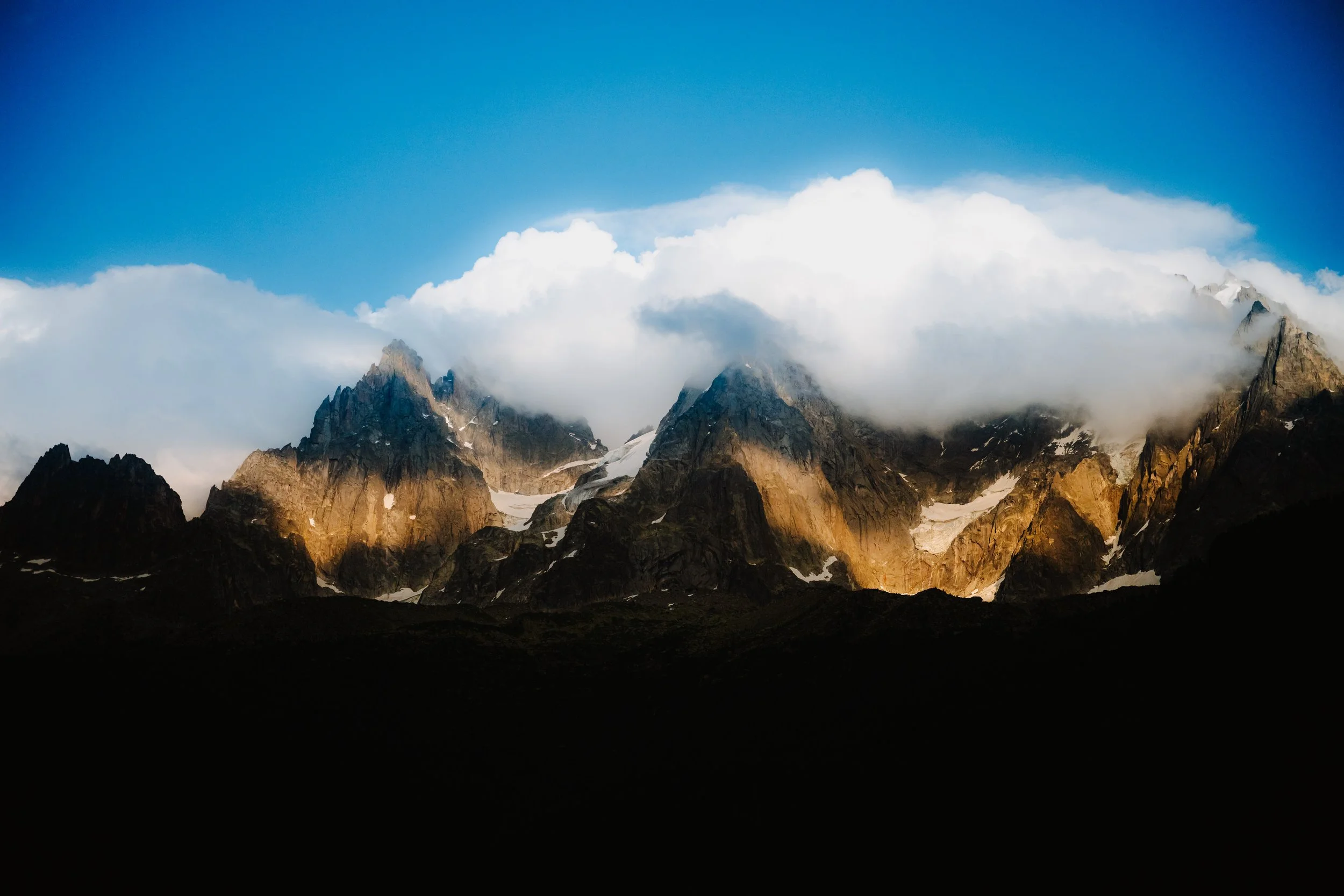Mountain range with rocky peaks partially covered by clouds, under a blue sky.