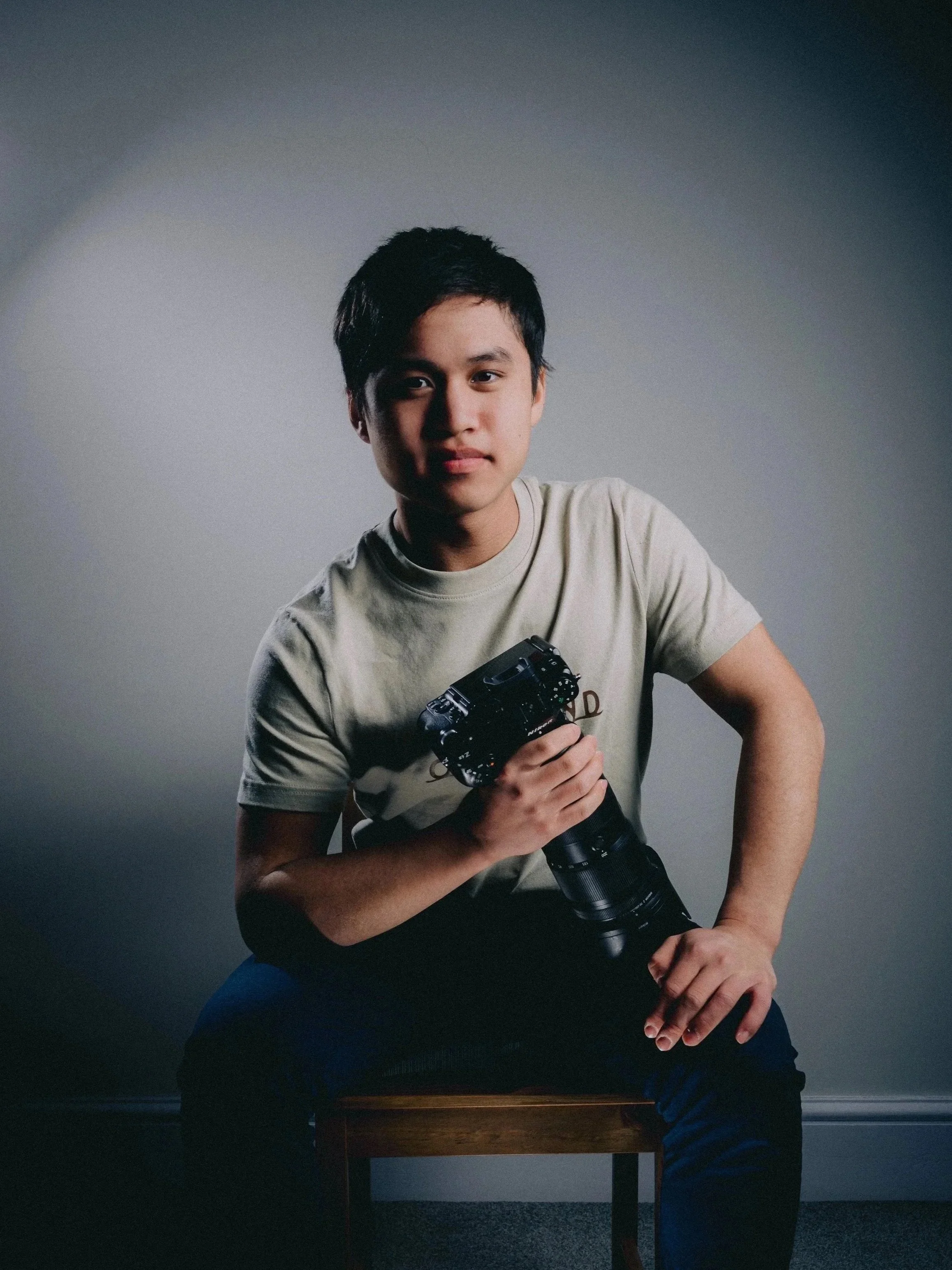 A young man sitting on a wooden chair holding a professional camera with a large lens, wearing a beige t-shirt, against a plain gray background.