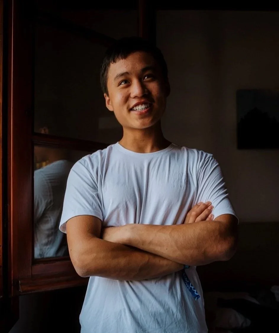 A young man with short dark hair wearing a white t-shirt stands with arms crossed, smiling in a dimly lit room.