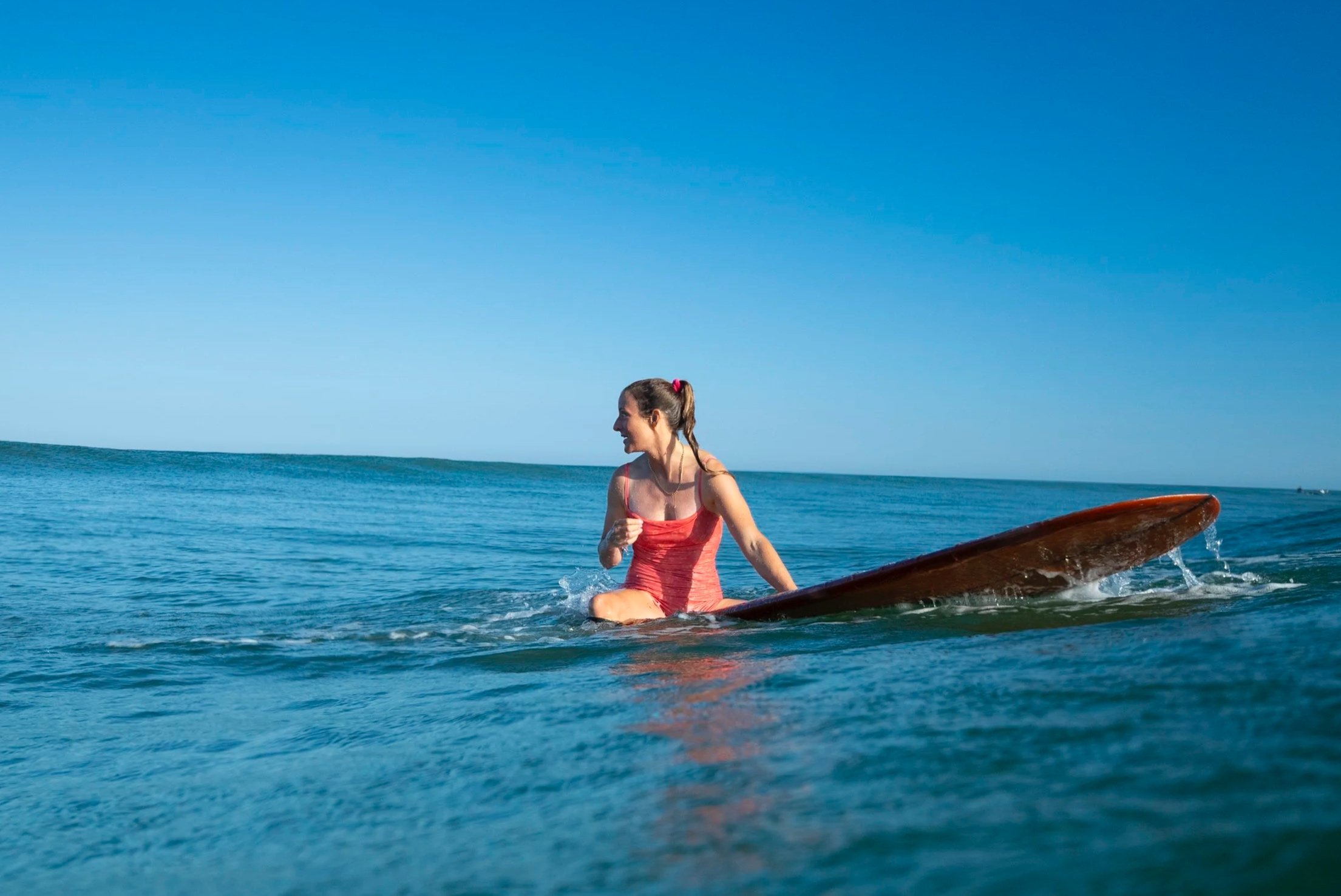 A woman in a pink swimsuit sitting on a surfboard in the ocean with a clear blue sky above.