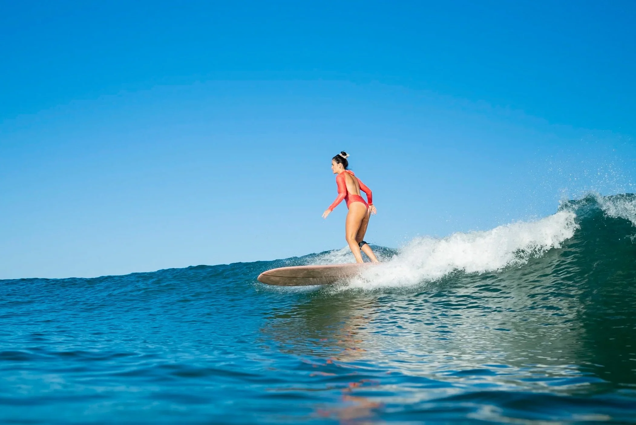A woman in a pink swimsuit surfing on a wave under a clear blue sky.