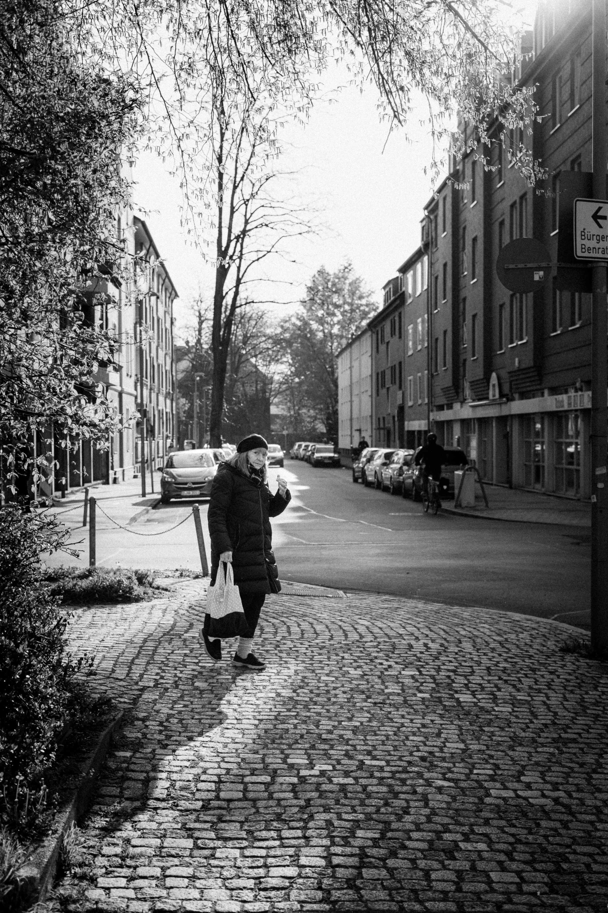 Black and white street photograph of a woman walking along a cobblestone sidewalk in Düsseldorf, Germany. The morning light casts soft shadows, highlighting the serene residential street lined with parked cars and traditional buildings.
