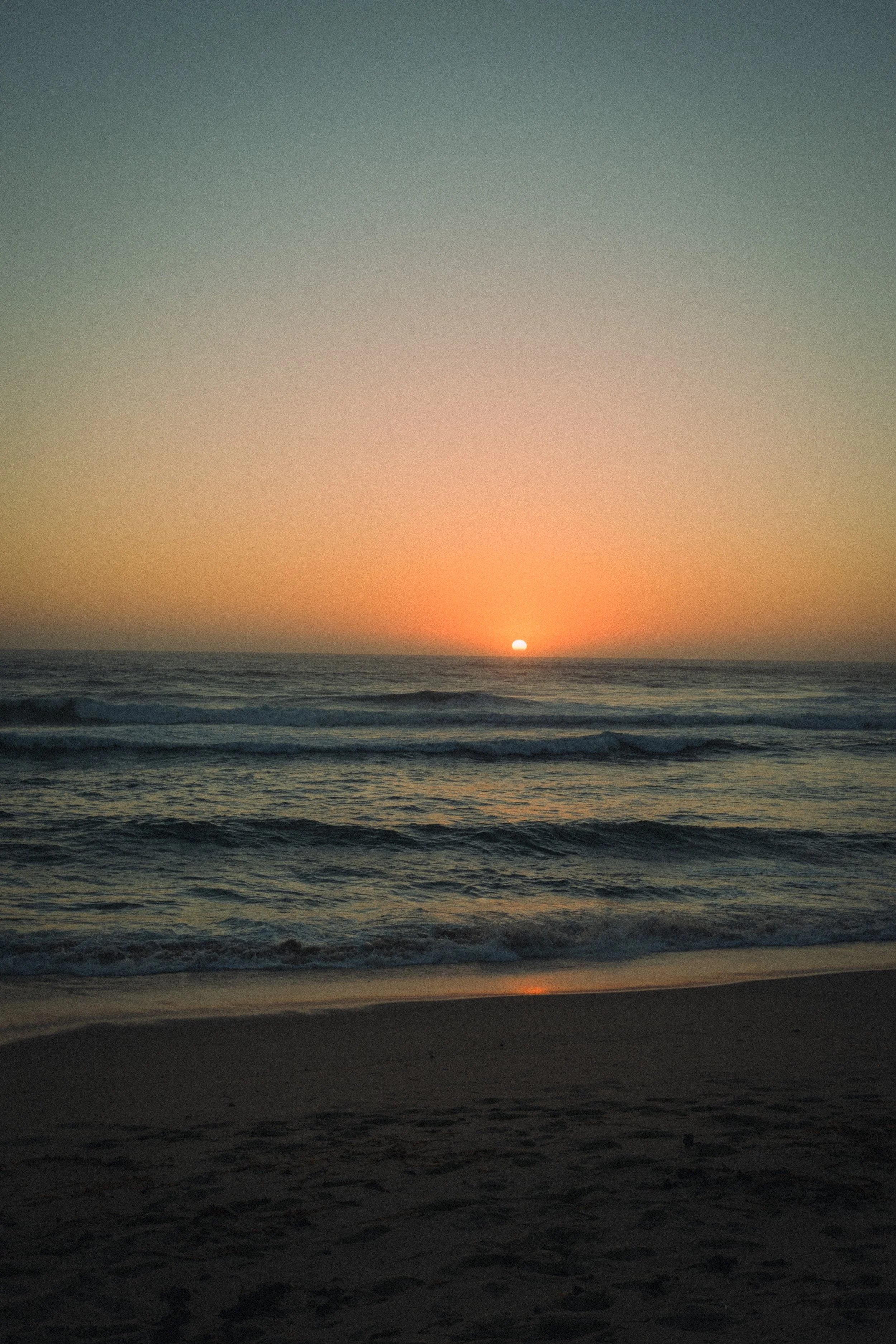 Fine art landscape photograph of a sunset in Ericeira, Portugal. In the foreground the sand, followed by the cold sea and the orange sun in the horizon with the orange/blue sky on top.