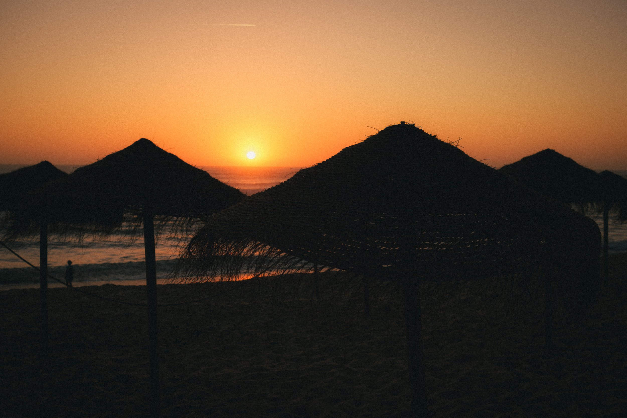 Fine art travel photography of a serene sunset on the beach in Ericeira, Portugal, featuring silhouetted straw parasols and golden hues reflecting on the ocean.