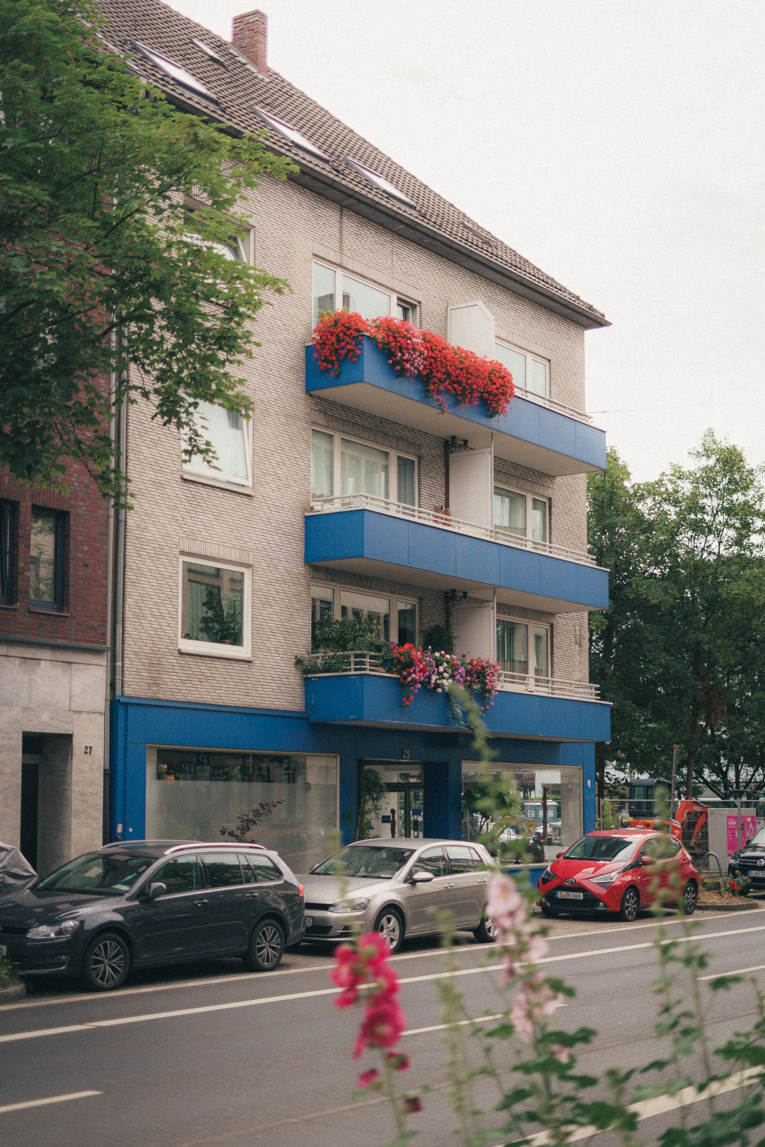 Fine art photograph of a building with blue balconies that contrast with the red and pink flowers on them, in Düsseldorf, Germany.