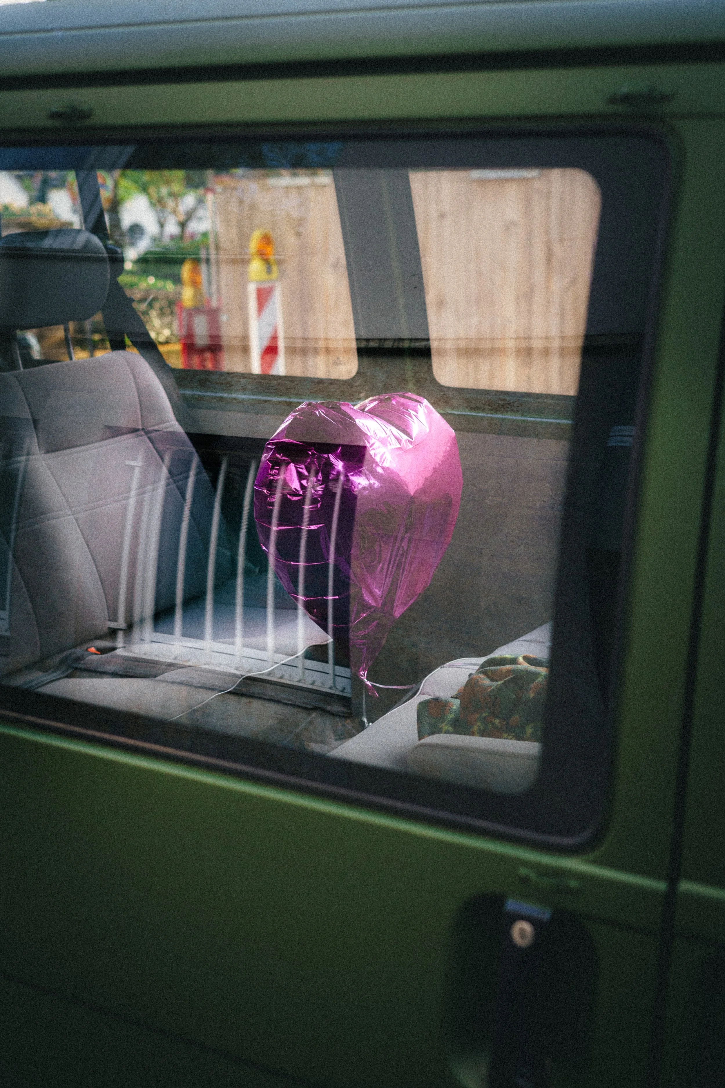 A vibrant pink heart-shaped balloon rests inside a green van, seen through the window. The reflection on the glass subtly blends exterior elements with the interior, creating a layered and intriguing visual composition in Düsseldorf, Germany.