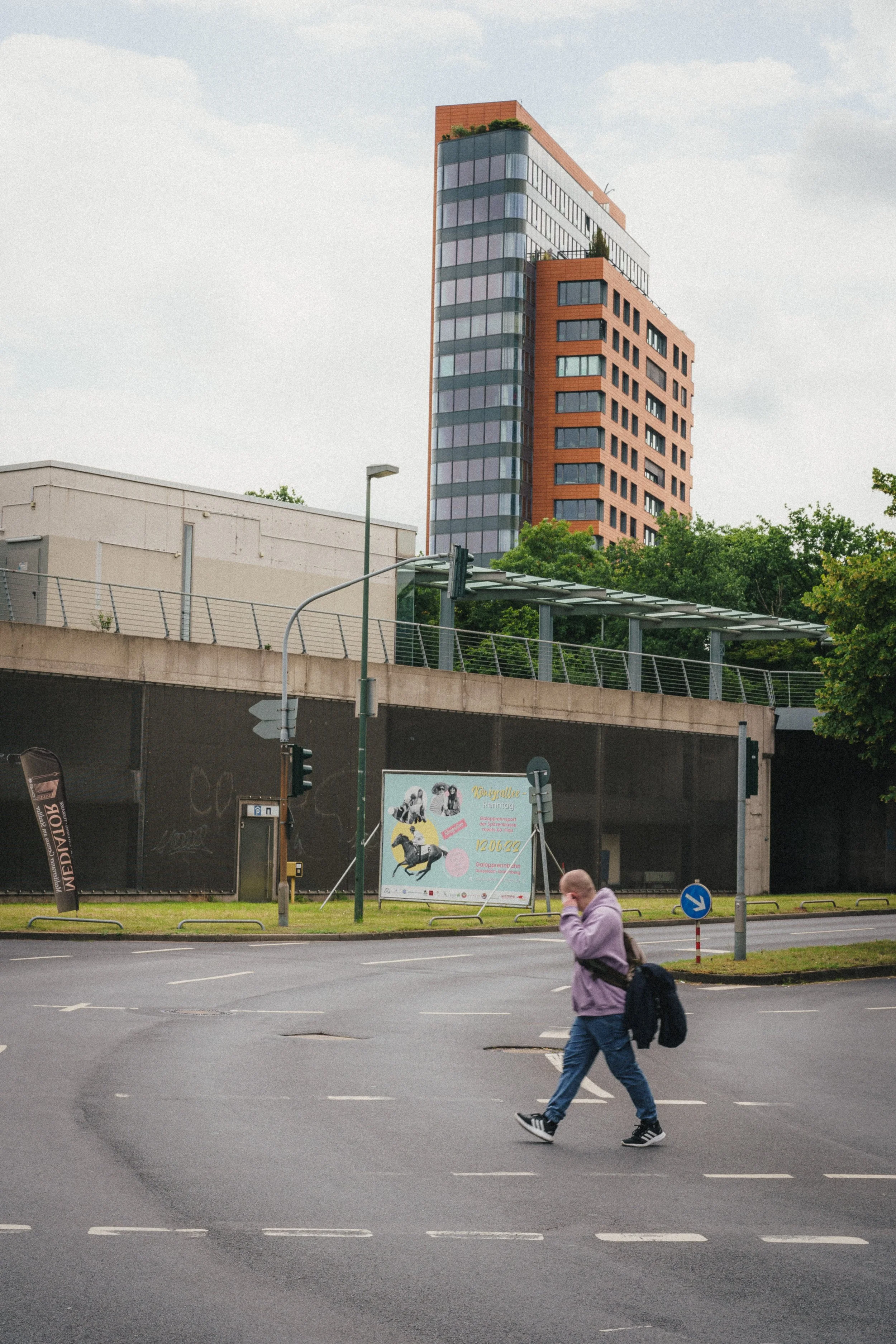 A pedestrian in a purple hoodie and jeans walks across a wet intersection in Düsseldorf. Behind him, an orange and gray modern high-rise building stands tall near an overpass, with advertisements and greenery in the background.