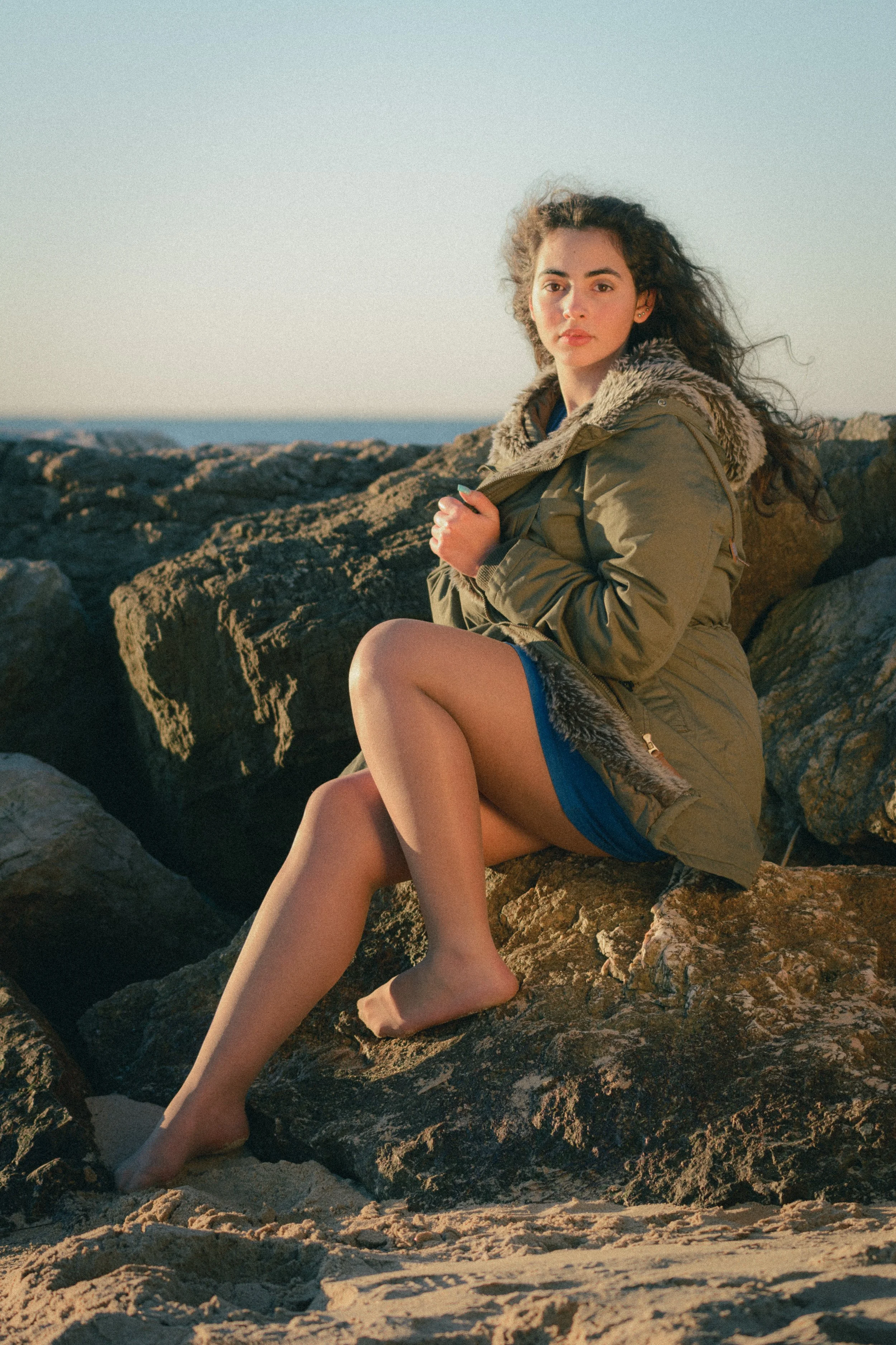 Portrait of Inês Chalupa in a beach in Portugal in Golden hour, wearing a blue dress and a green coat looking at the camera, sitting in the rocks grabbing her coat, with the blue sky behind her.