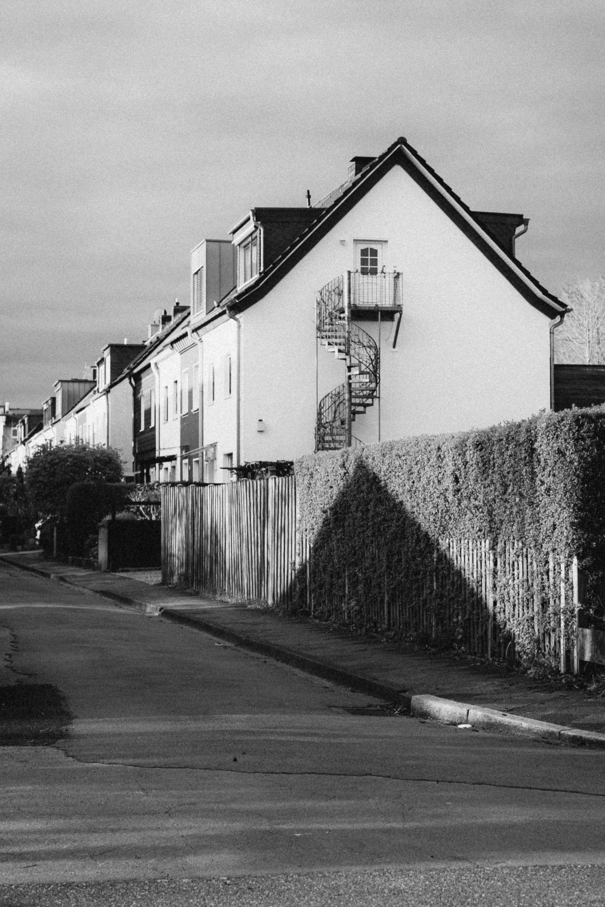 Black and white photograph of a residential street in Düsseldorf, Germany, featuring a white house with a spiral staircase on its side. The composition captures the play of light and shadow on the building and the neatly trimmed hedges lining the qui