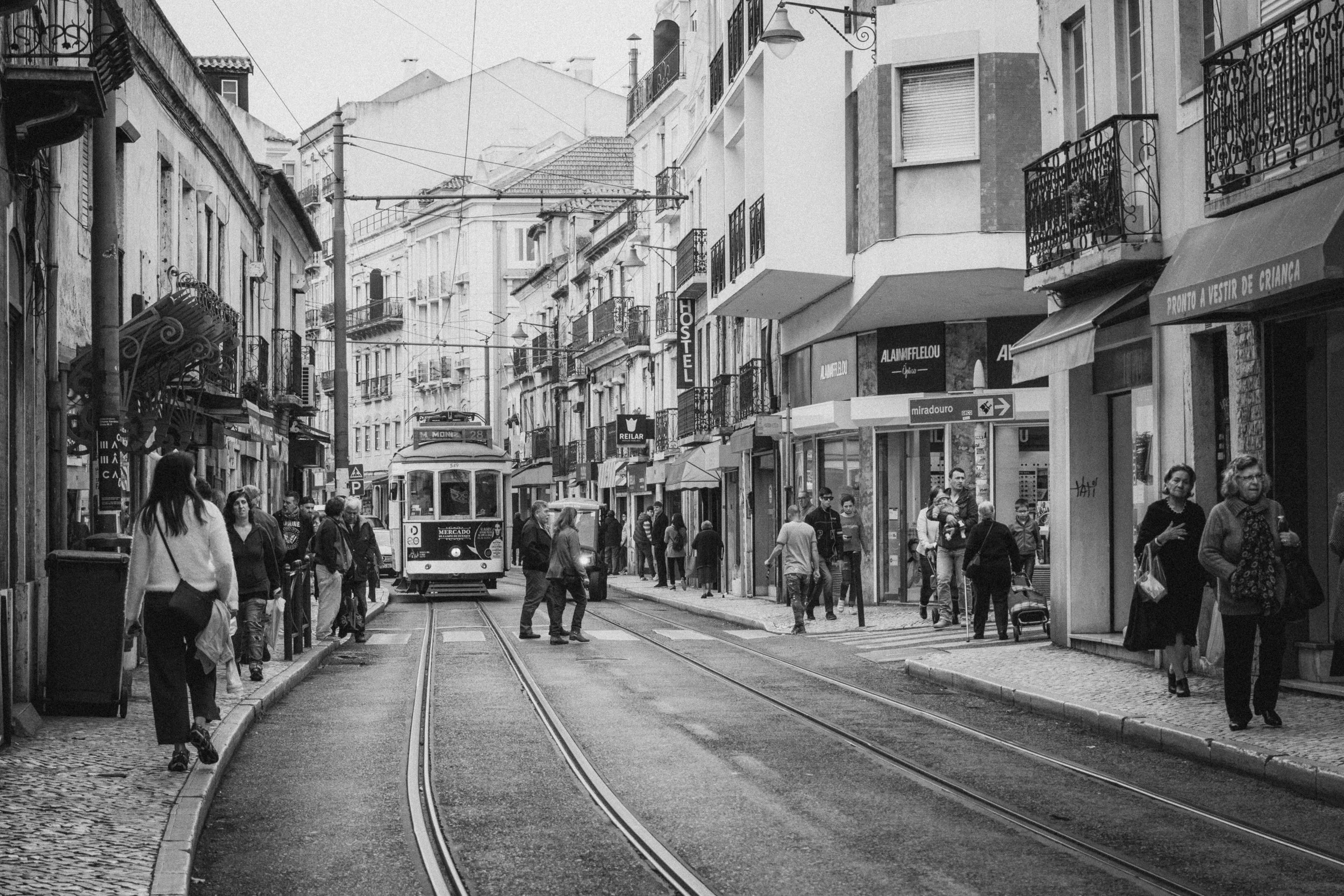 A bustling street in Lisbon, Portugal, featuring the iconic tram 28 moving along tracks surrounded by historic buildings and storefronts. Pedestrians fill the sidewalks, adding life to the scene, while the black-and-white tone enhances the timeless c