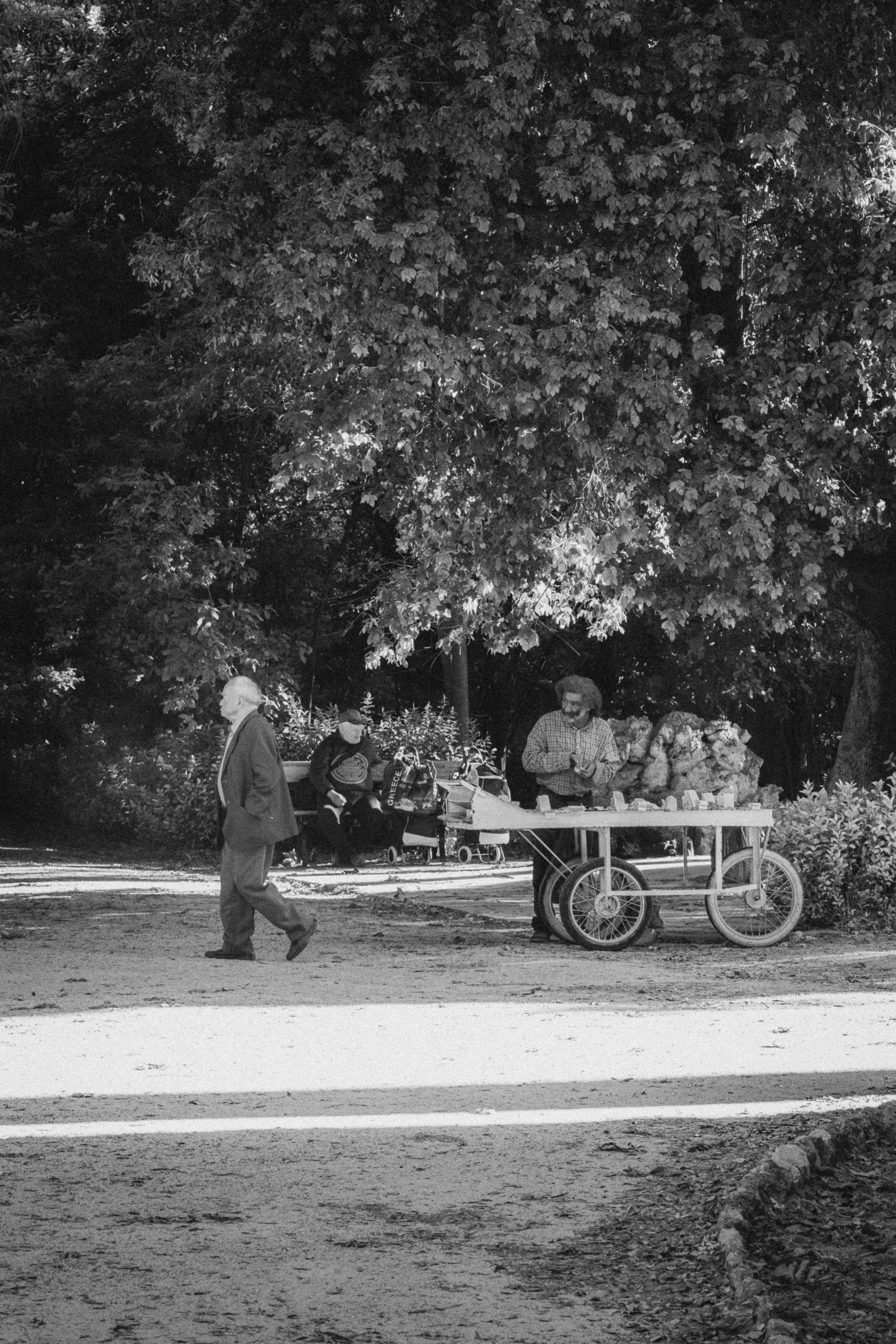 A black-and-white photograph taken in Athens, Greece, featuring a man walking through a park while another man stands by a cart selling goods, possibly fruits or vegetables. In the background, two elderly individuals are seated on a bench under the s