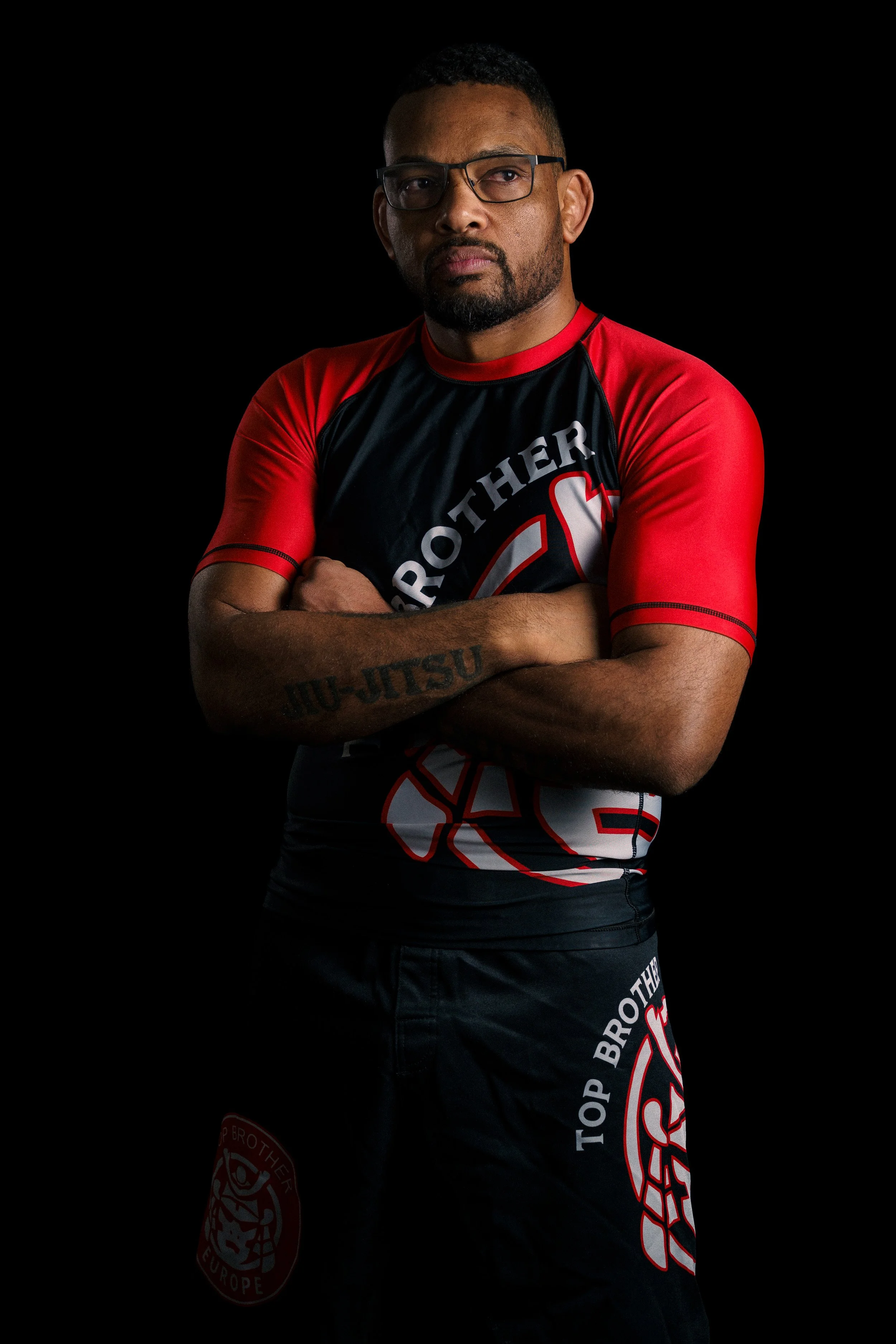 Studio portrait of a Brazilian Jiu-Jitsu athlete, Alex Malheiro from Top Brother, in nogi clothes, standing confidently in a training hall, crossed arms.