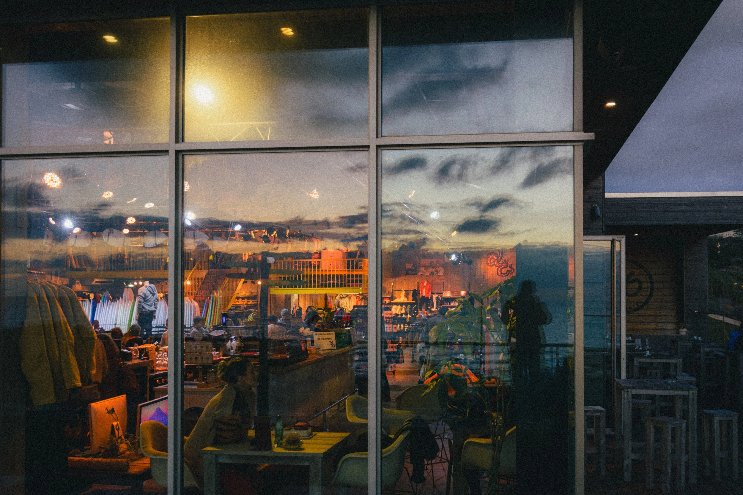 An atmospheric evening photograph showing the interior of a surf shop and cafe in Ericeira, Portugal, viewed through large glass windows. The vibrant interior lighting contrasts with the reflection of a dramatic sunset sky on the glass. Surfboards, c