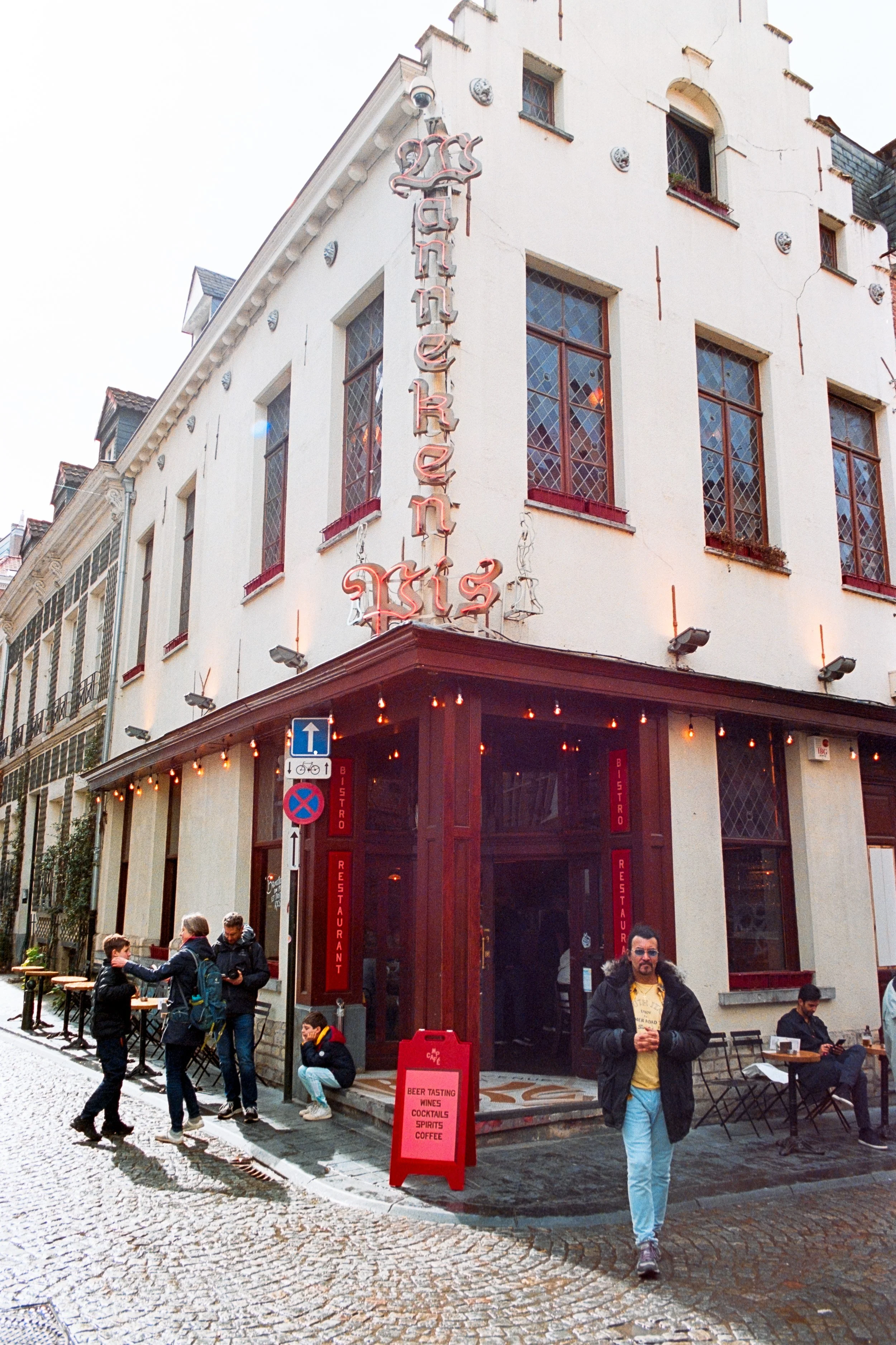 A lively street scene in Brussels featuring Heitor Silva walking with a cup in hand outside Manneken Pis Bistro. The charming white building is adorned with classic red signage and lit string lights, surrounded by people enjoying the cobblestone stre