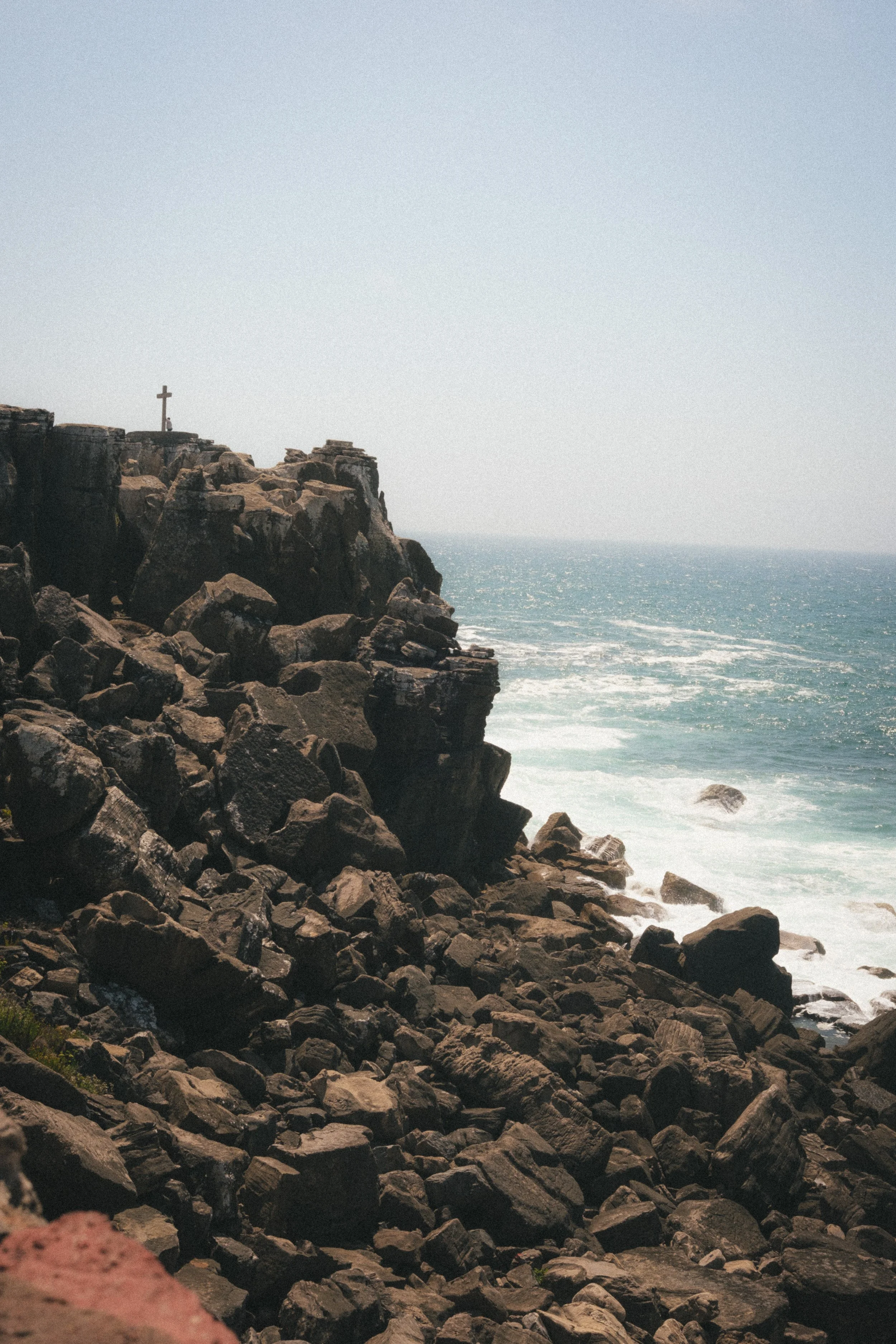 Fine art travel photography of rocky cliffs in Peniche, Portugal, with a cross perched atop the rugged landscape and the Atlantic Ocean in the background.