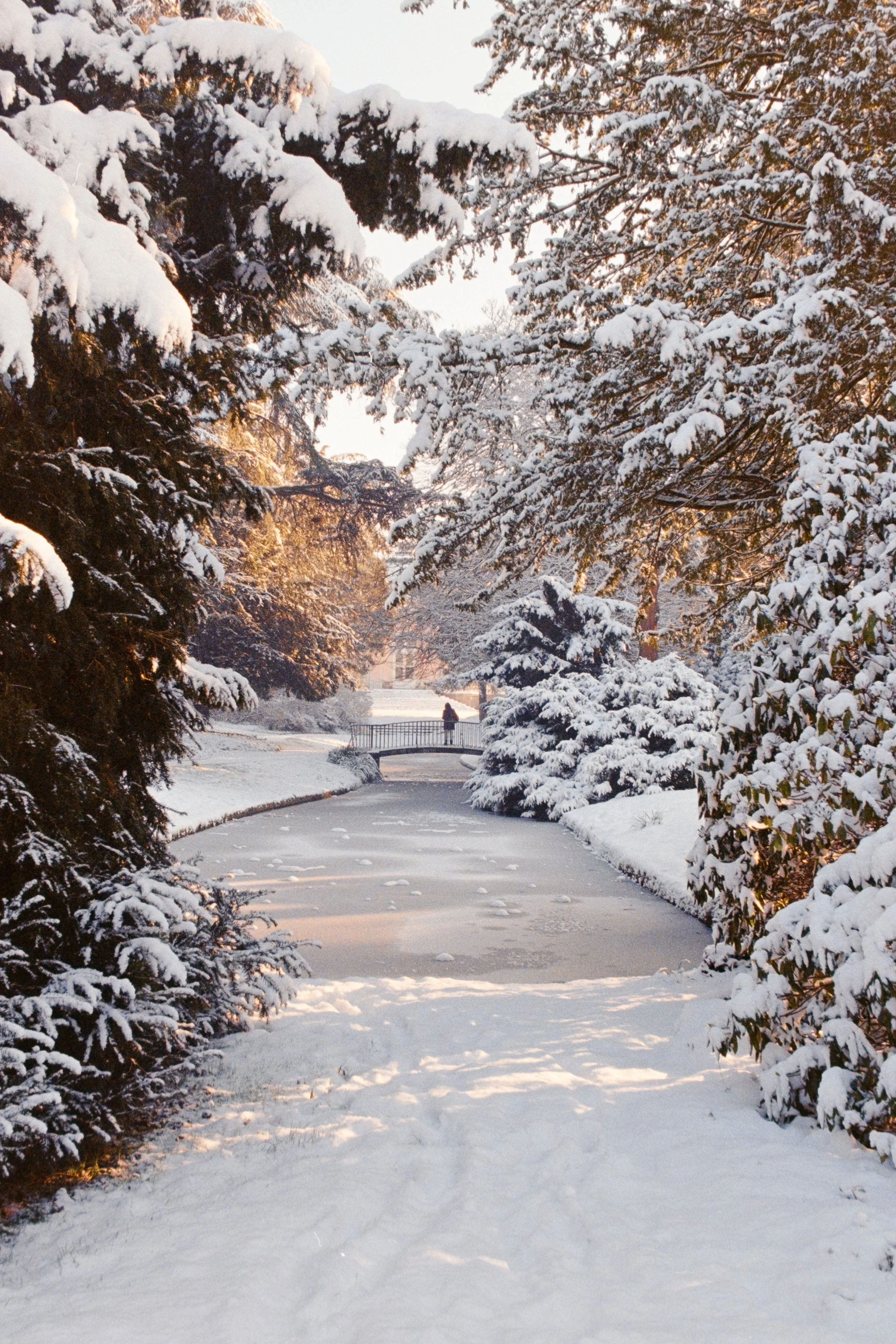 Fine art travel photography of a snowy landscape in Schloss Benrath Park, Düsseldorf, Germany, featuring a frozen stream and a pedestrian bridge framed by snow-covered trees.