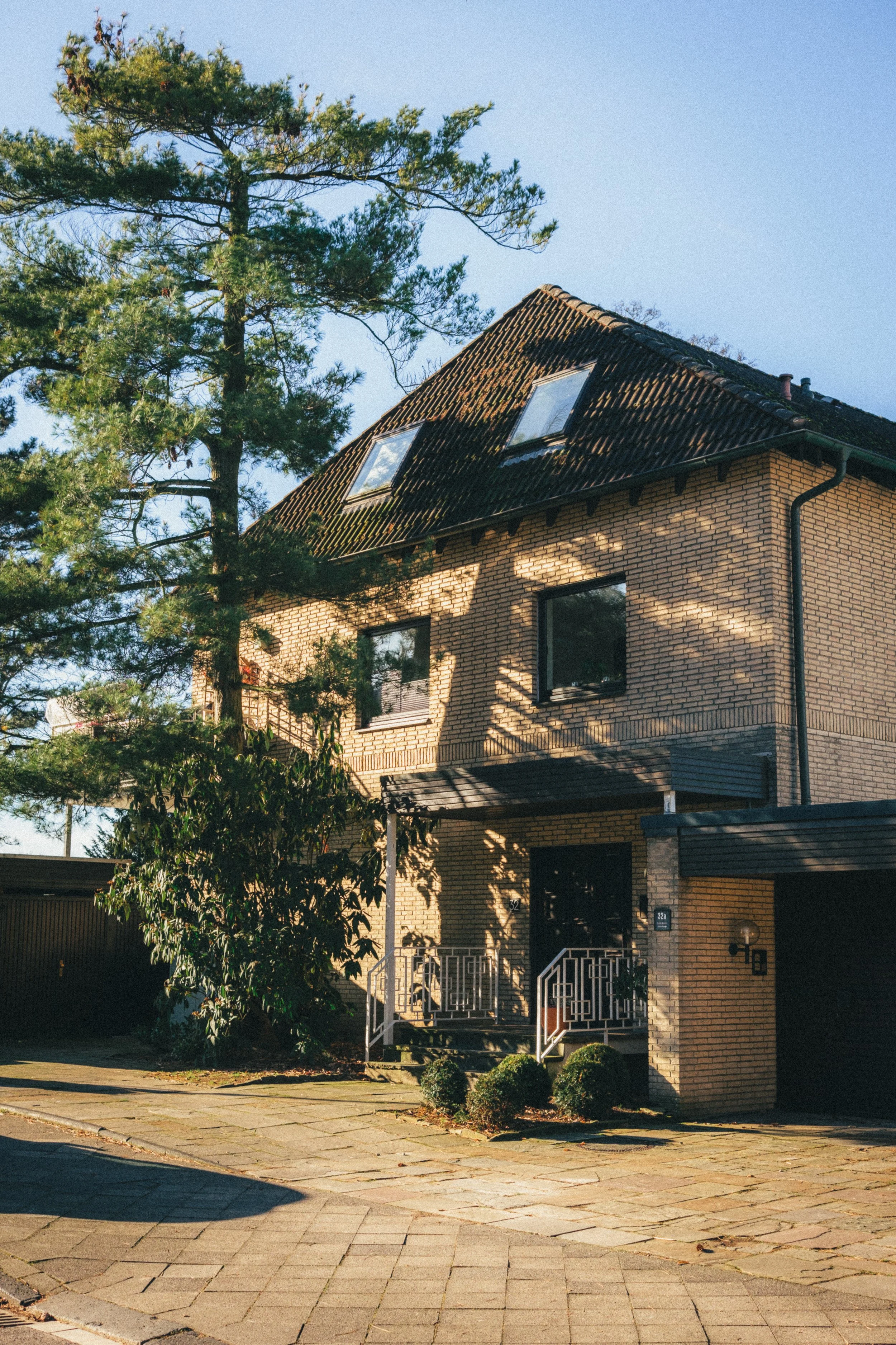 Warm, sunlit photograph of a charming brick house in Düsseldorf, Germany. The house features a sloped tiled roof with skylights, a lush tree in the foreground, and a neatly paved driveway, evoking a tranquil suburban ambiance.