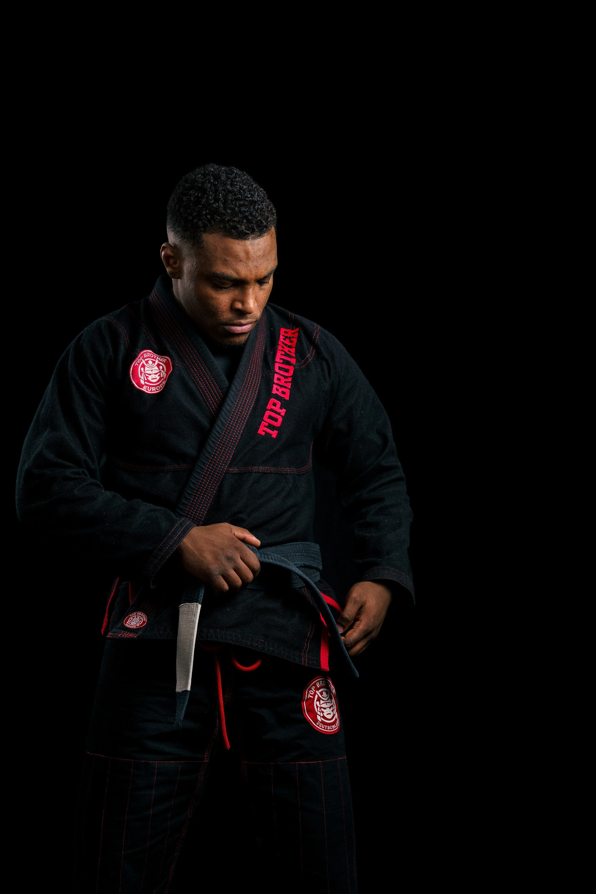 Studio portrait of a BJJ athlete, Renan Malheiro, tying his black belt, focusing on the hands, wearing a black gi.