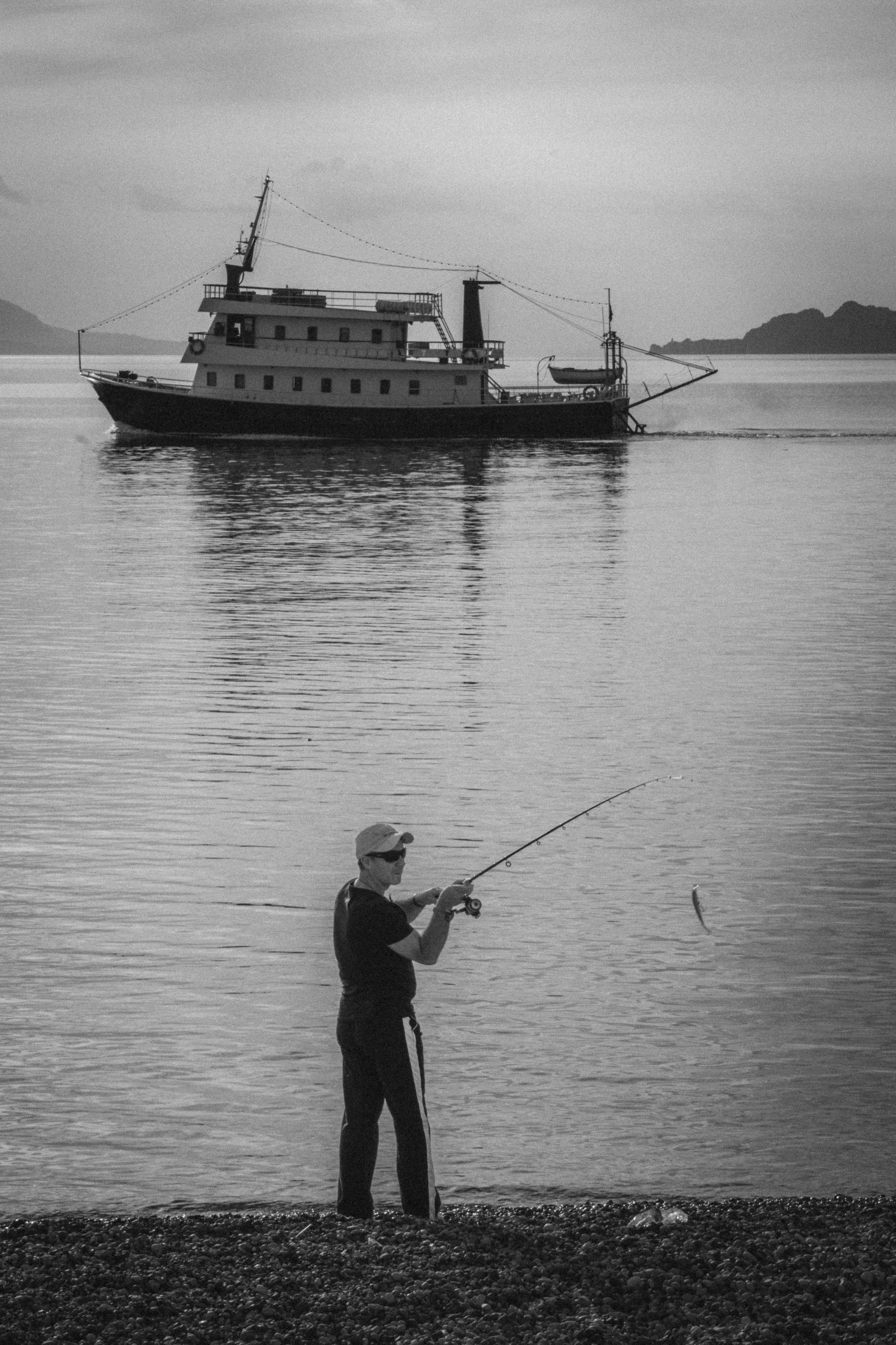 A serene black-and-white image of a man fishing along the pebbled shore in Loutraki, Greece. In the background, a vintage boat sails peacefully on calm waters, with distant hills faintly visible under the soft, diffused light. The captured fish mid-a