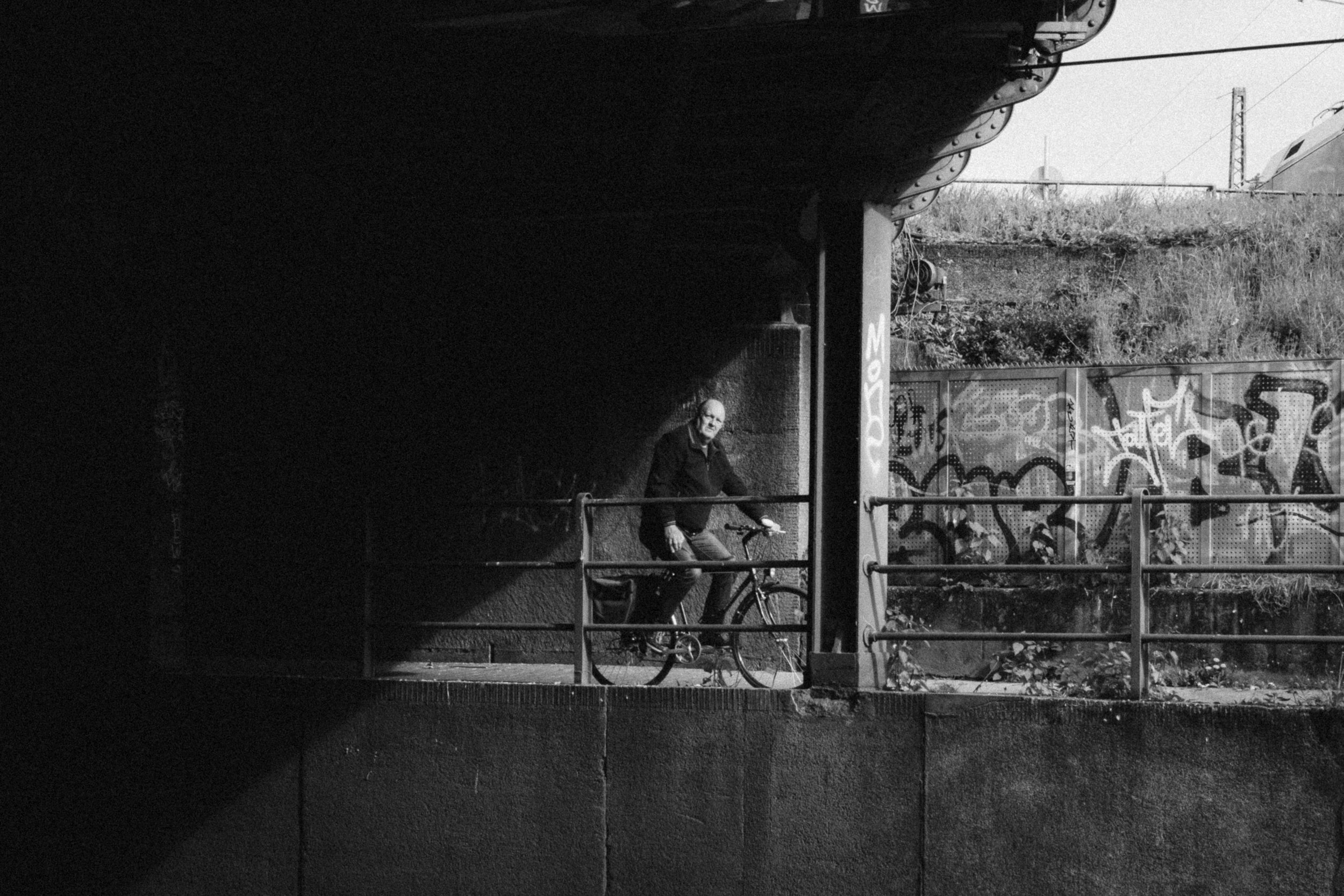 Black and white photograph of a man on a bicycle under a bridge in Düsseldorf, Germany. The interplay of light and shadow highlights the subject against a backdrop of graffiti-covered walls, adding an urban and contemplative feel to the scene.