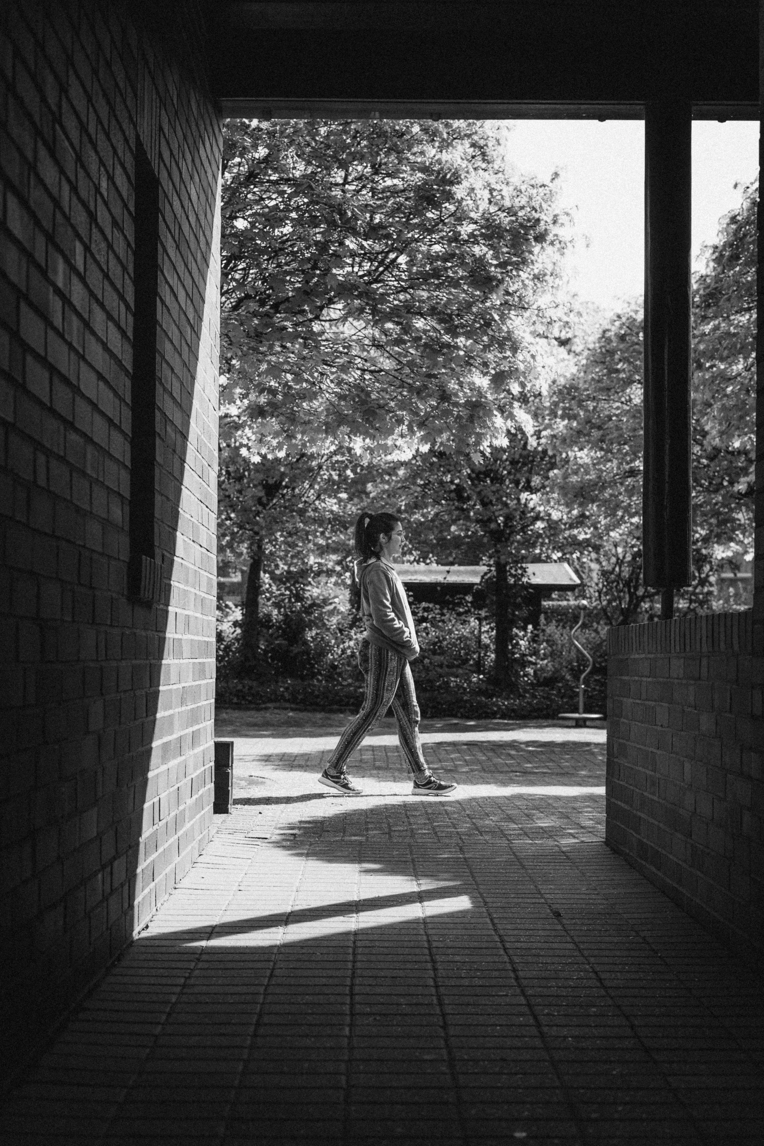Black and white photograph of a young woman walking past a brick corridor in Düsseldorf, Germany. The interplay of light and shadow creates a framed view of the subject against a backdrop of trees, blending urban and natural elements.