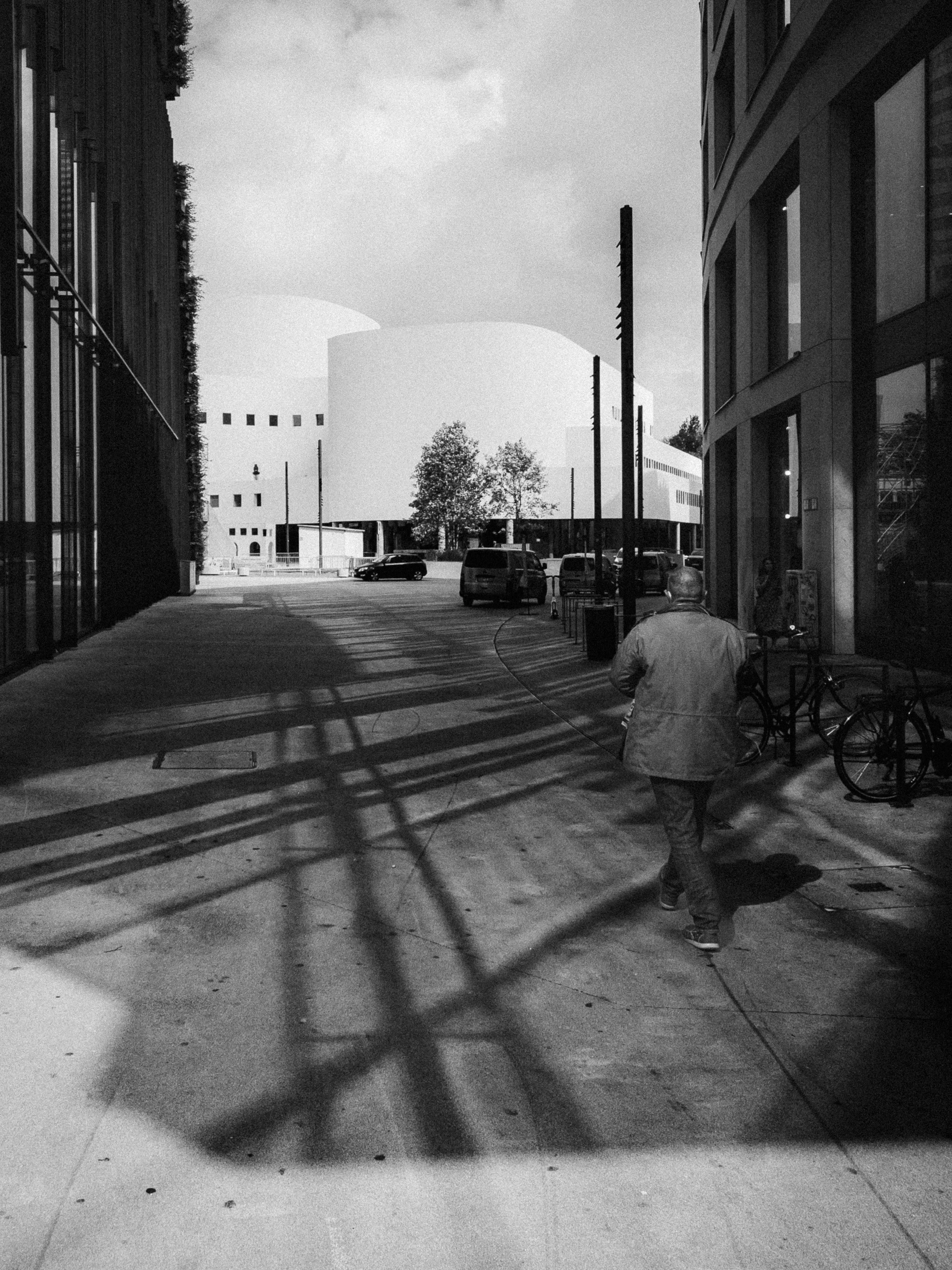 A monochrome urban scene in Düsseldorf, Germany, showcasing modern architectural lines. A lone pedestrian walks along a pathway where dynamic shadows of a structural framework create intersecting patterns on the ground. Contemporary buildings frame t