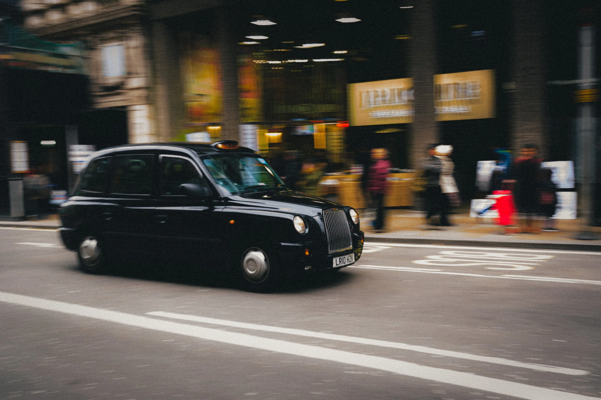 A black London taxi in motion on a busy street in London, UK. The iconic black cab stands out against the blurred backdrop of pedestrians and shop windows, capturing the dynamic and bustling atmosphere of the city.