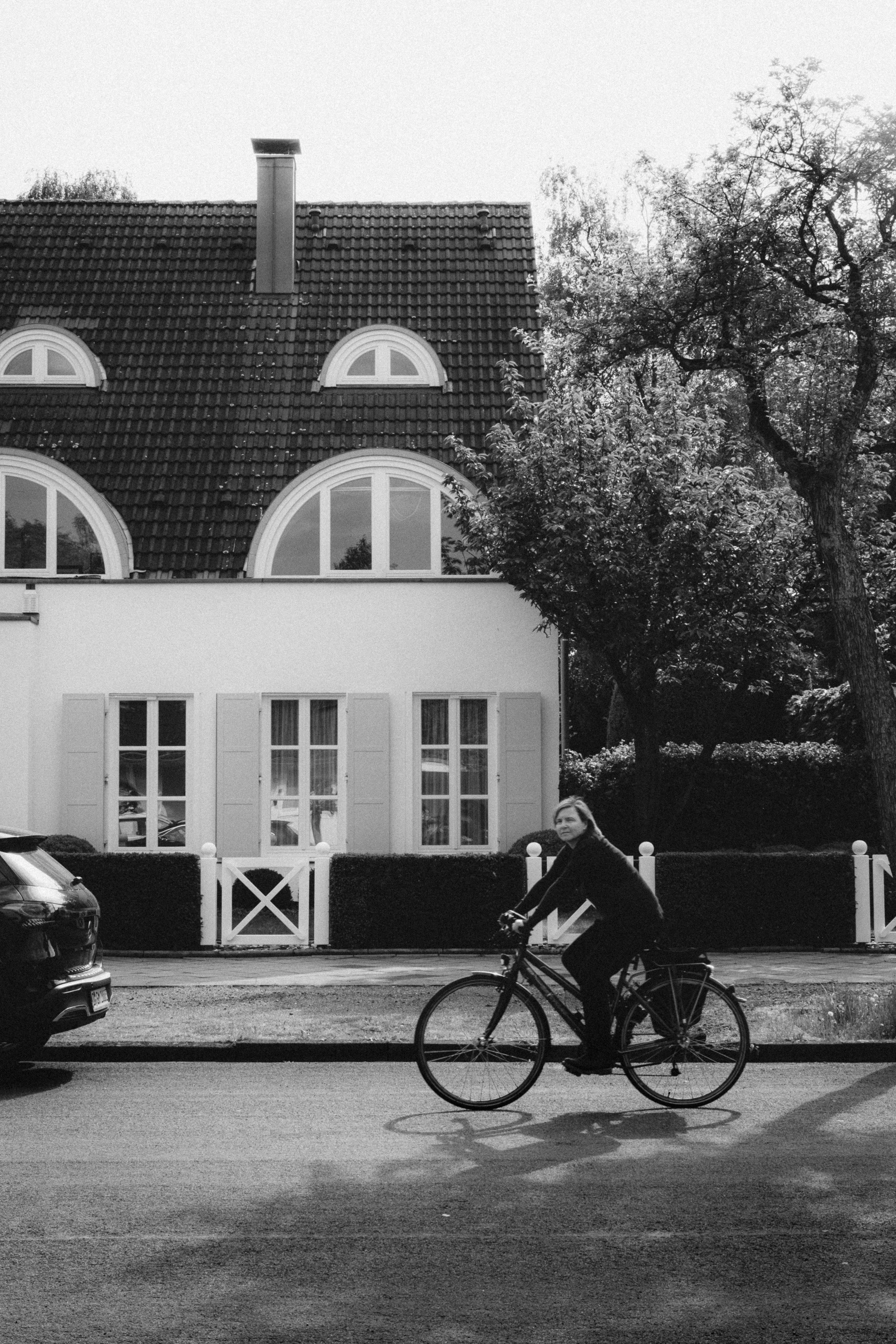 Fine art black and white street photograph of a woman cycling past a charming house with arched windows in Düsseldorf, Germany. The elegant architecture and lush greenery create a serene suburban scene, blending activity and tranquility.