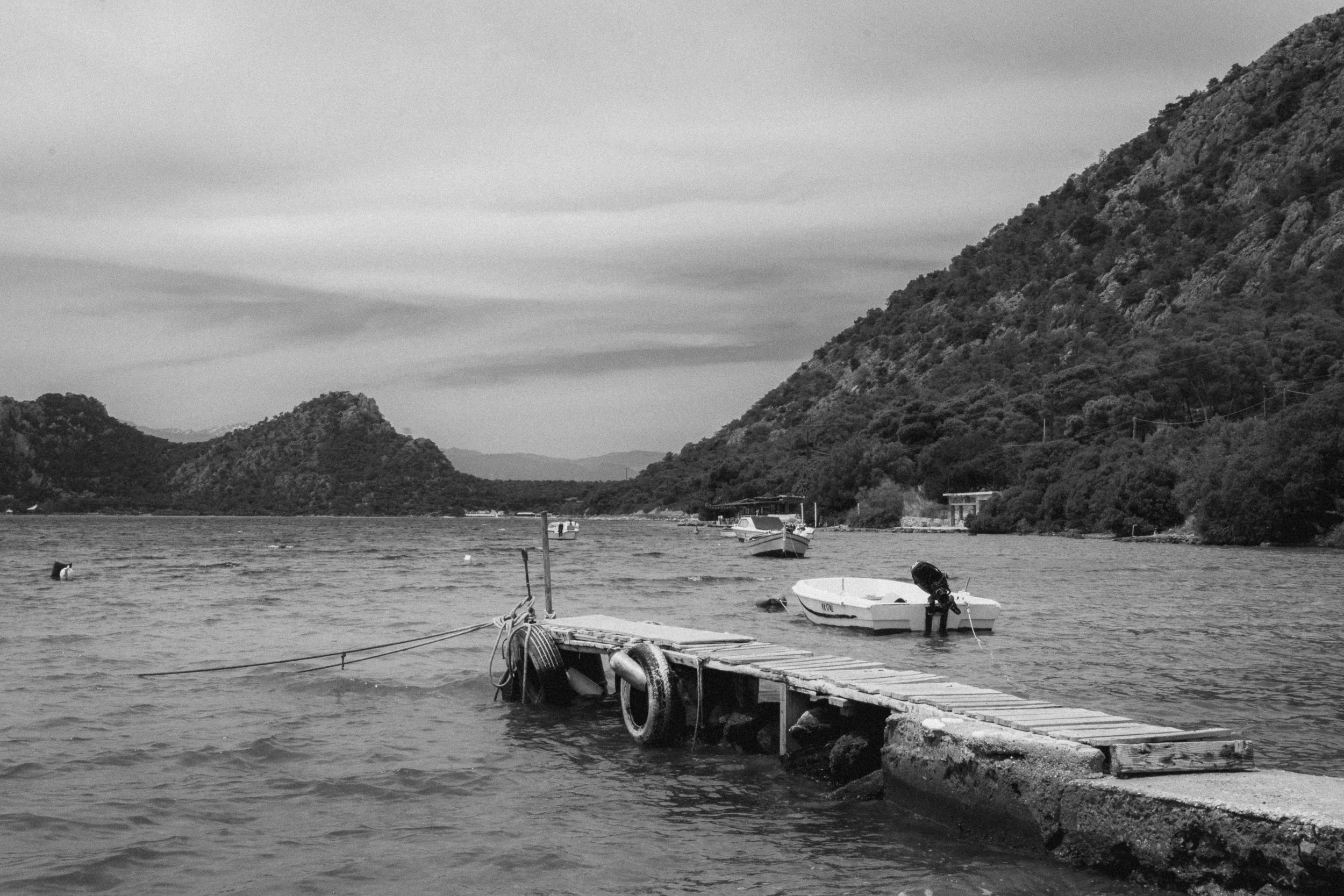 Fine art black and white landscape of a beach in Greece. A bridge stands across with both on the right, mountain on the horizon and a beautiful sky behind them.