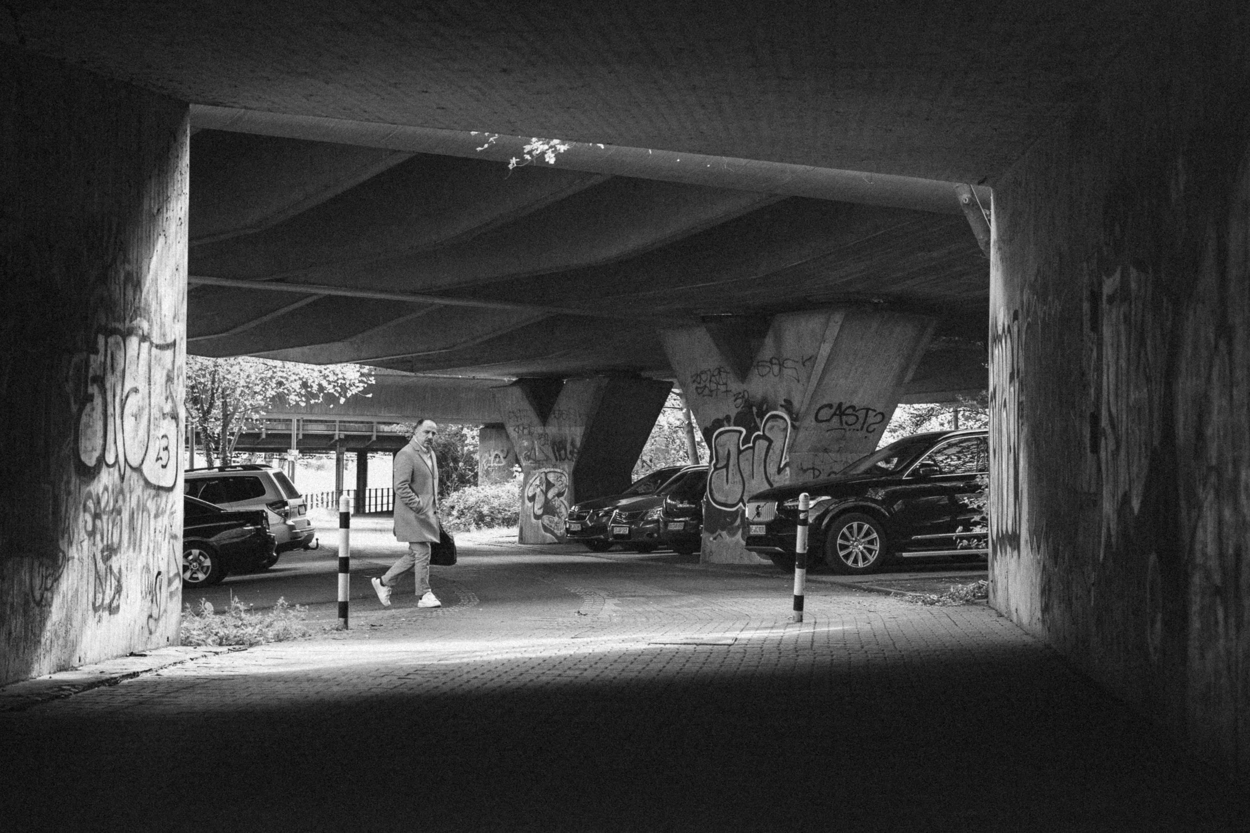 Fine art black and white street photograph of a man walking under a graffiti-covered overpass in Düsseldorf, Germany. The urban setting, with concrete pillars and parked cars, contrasts with the soft natural light filtering through, creating a gritty