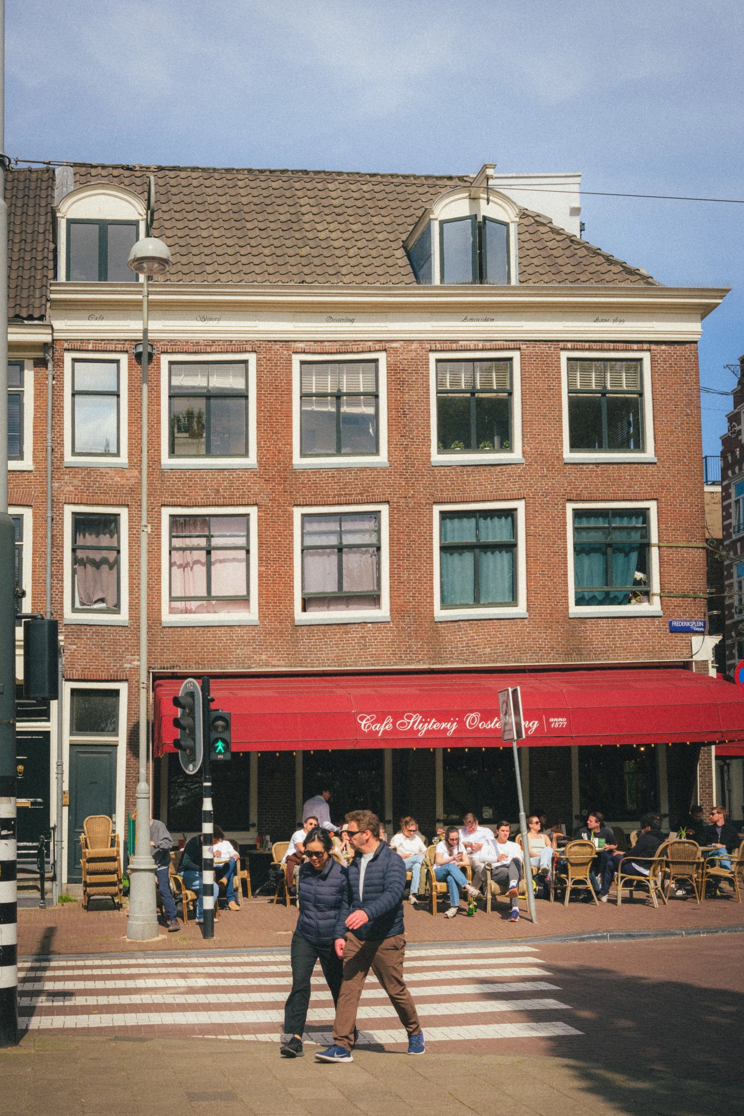 Café Slijterij Oosterling in Amsterdam, a historic red brick building with a vibrant red awning, captured on a sunny day with pedestrians crossing and outdoor seating. Fine art urban photography showcasing Dutch culture.