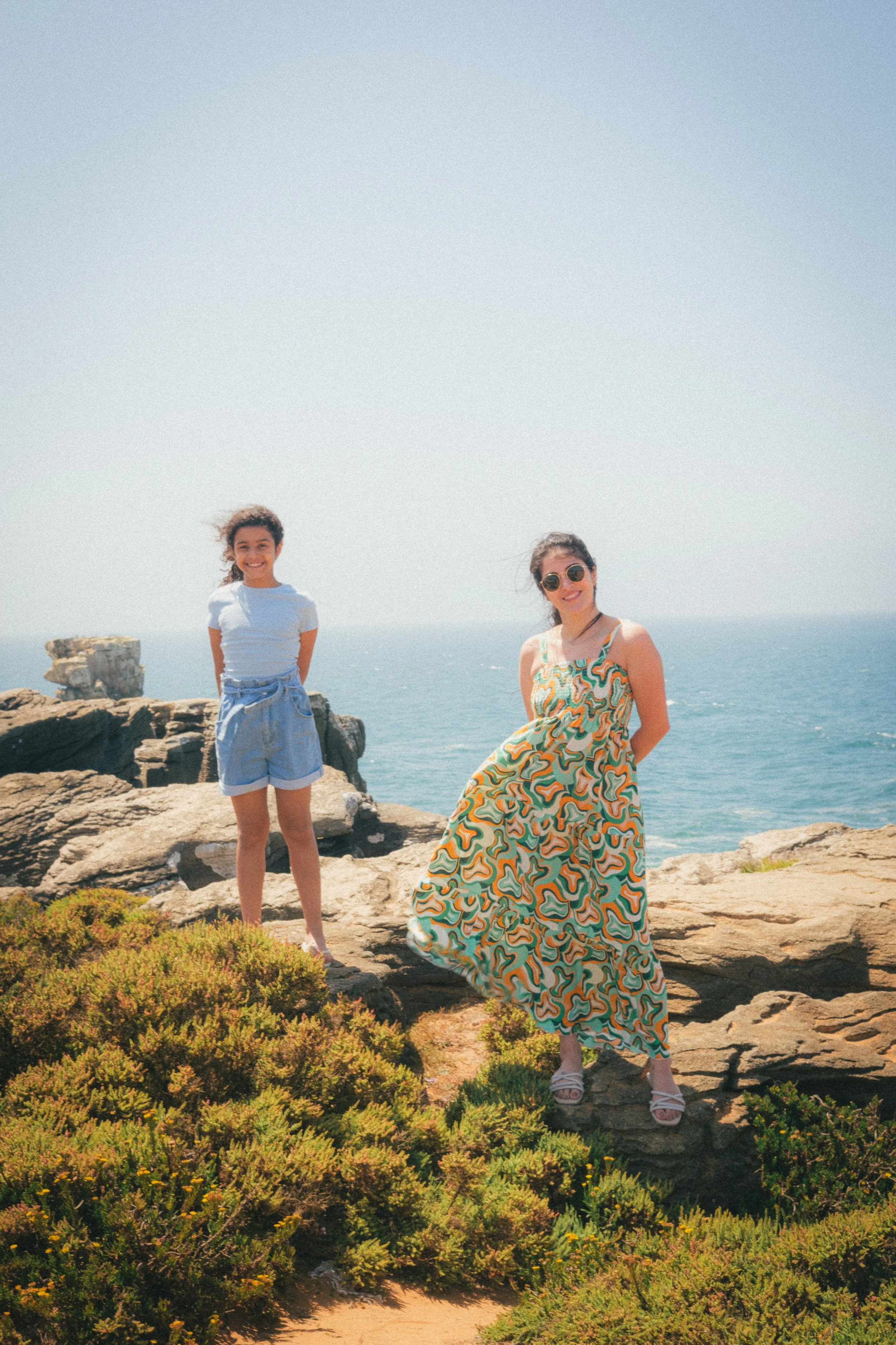 Fine art travel photography of a vibrant coastal scene in Peniche, Portugal, featuring two individuals standing on rocky cliffs overlooking the ocean, with lush greenery in the foreground.