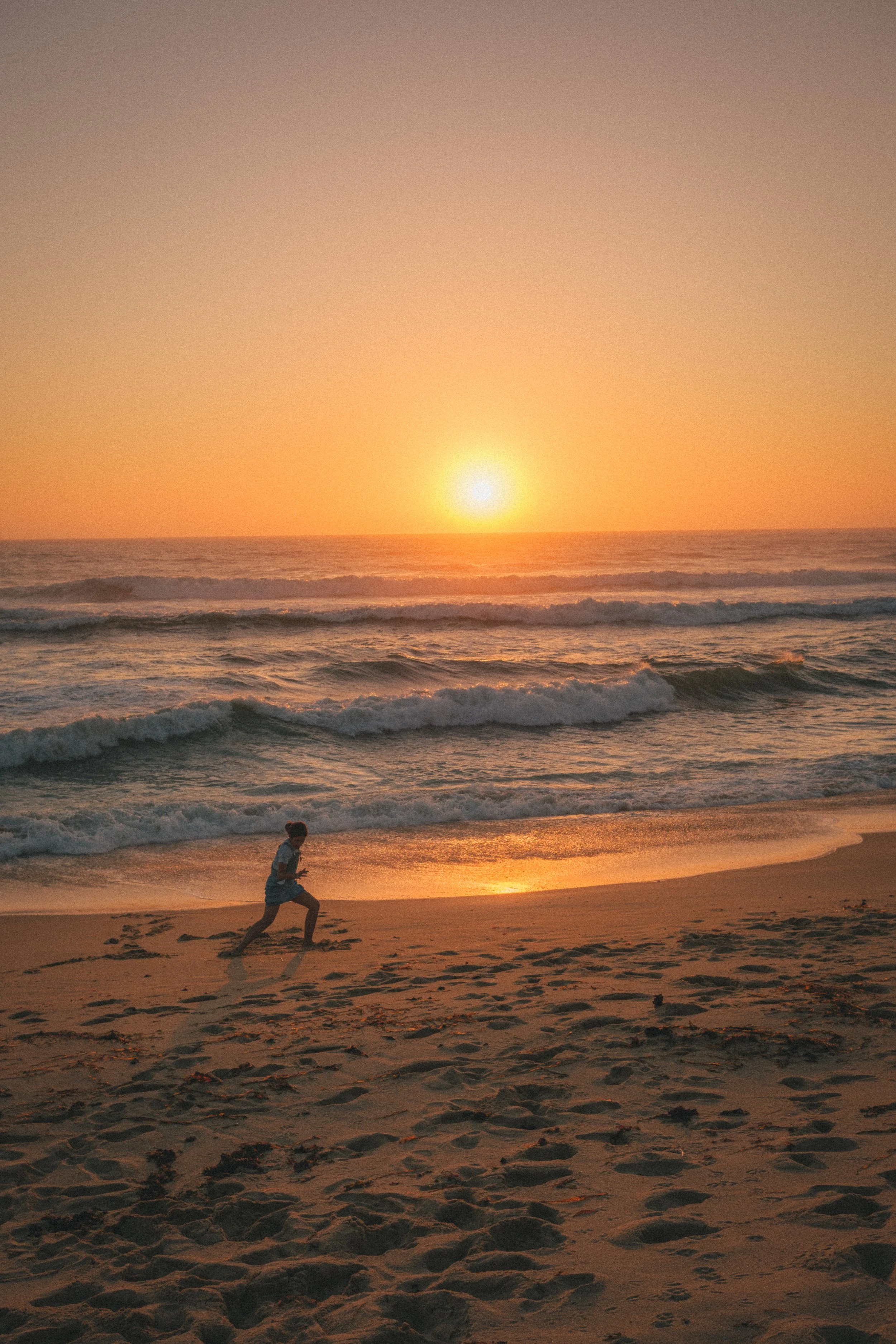 Fine art travel photography of a person jogging along the sandy beach during a golden sunset in Ericeira, Portugal, with ocean waves in the background.