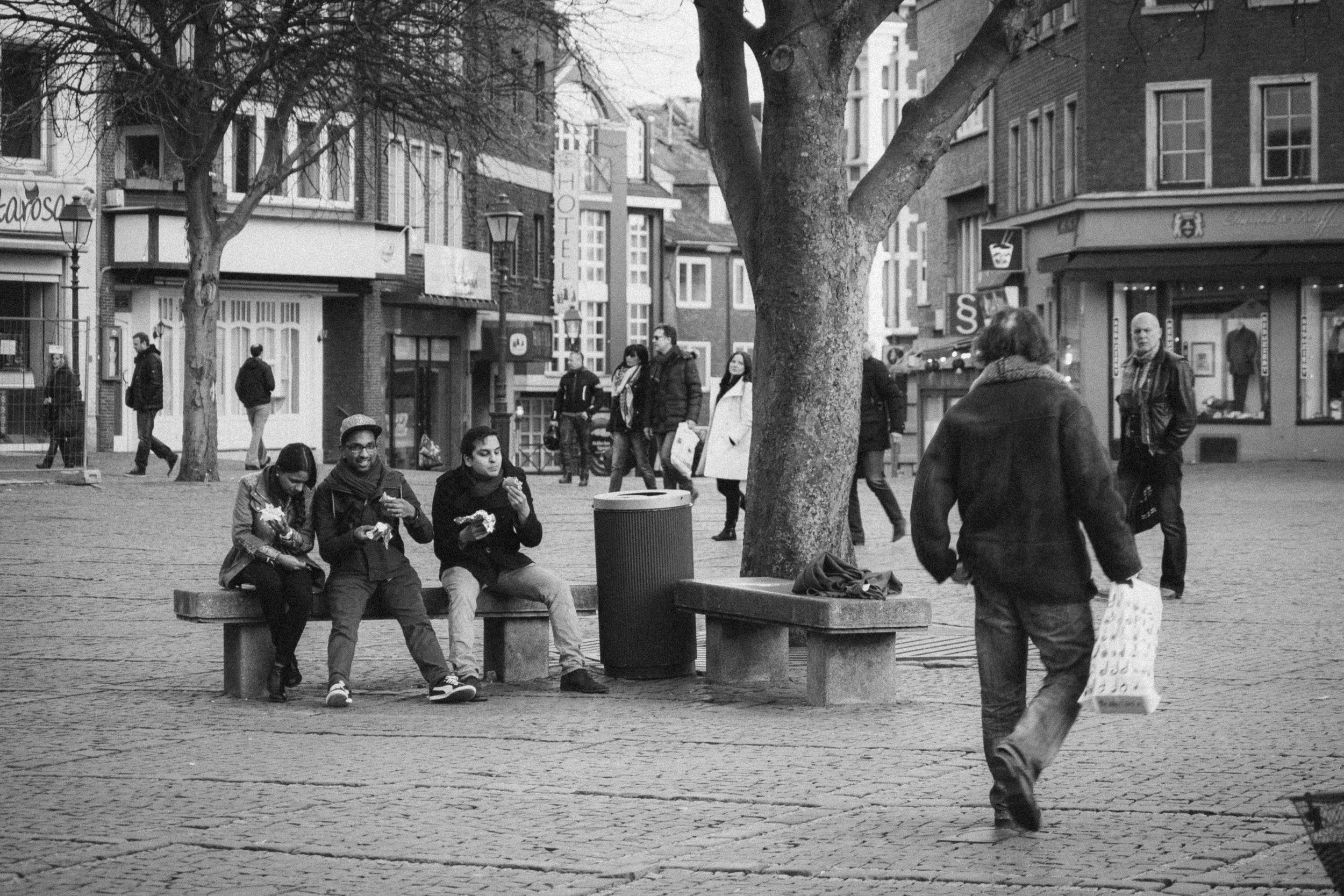 A black-and-white photograph taken in Aachen, Germany, capturing a lively urban scene. Three people sit on a bench in a cobblestone square, enjoying food and engaged in conversation. Behind them, pedestrians walk past shops and historic buildings, wh