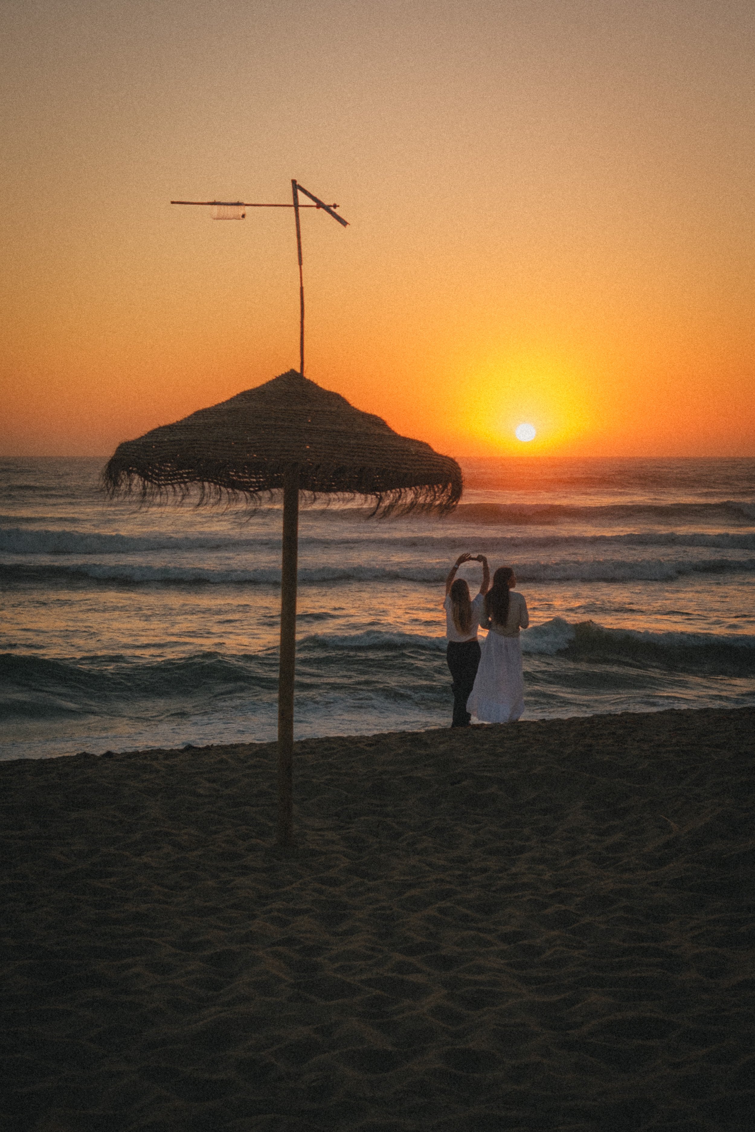 Fine art travel photography of a romantic sunset on the beach in Ericeira, Portugal, with two people silhouetted near the ocean, framed by a straw parasol and glowing orange sky.