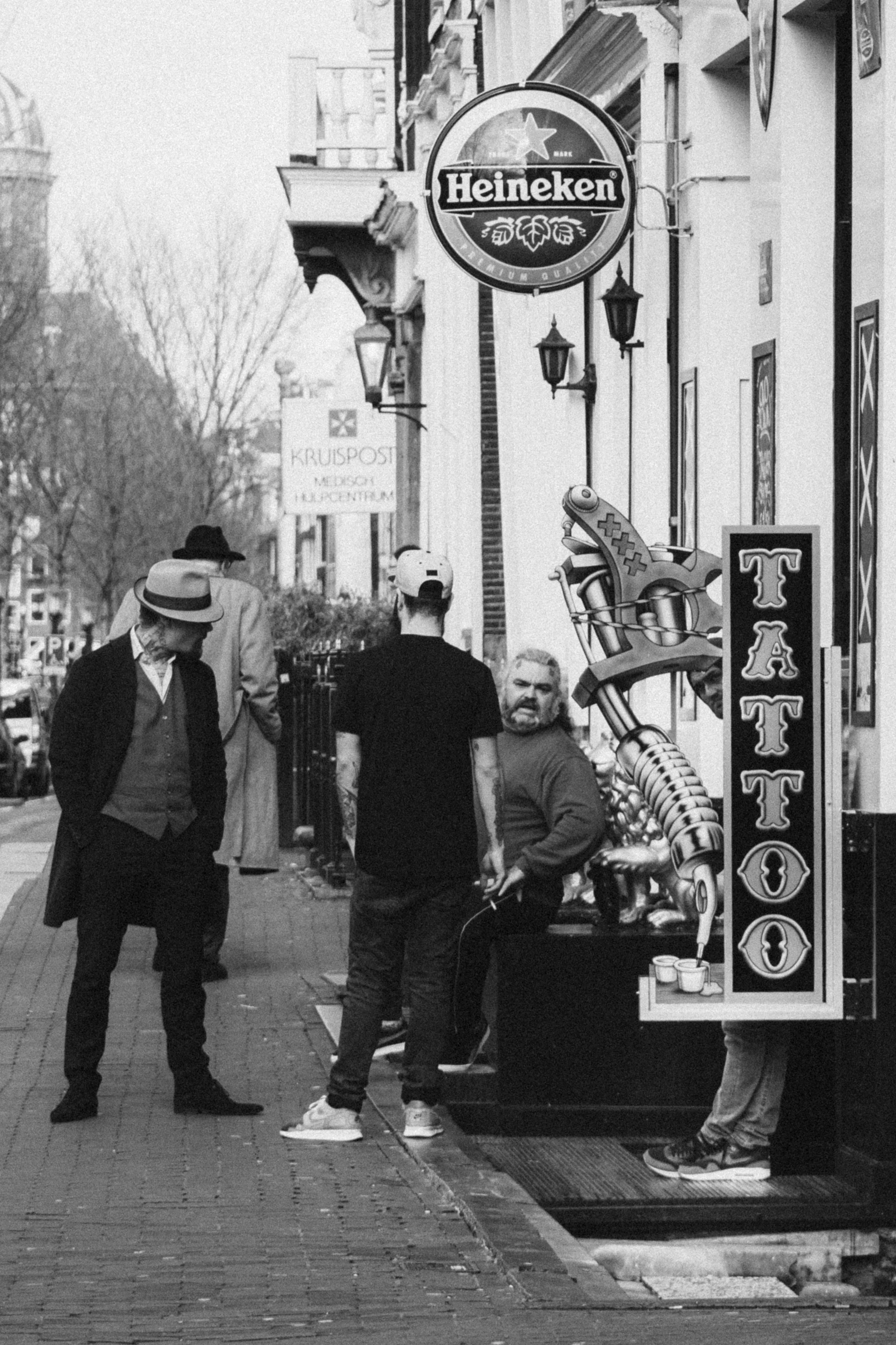 A candid black-and-white street scene in Amsterdam, featuring three men near a tattoo parlor. One man leans casually against a Heineken sign, while another looks over his shoulder. A large tattoo machine sign adds a striking element to the storefront