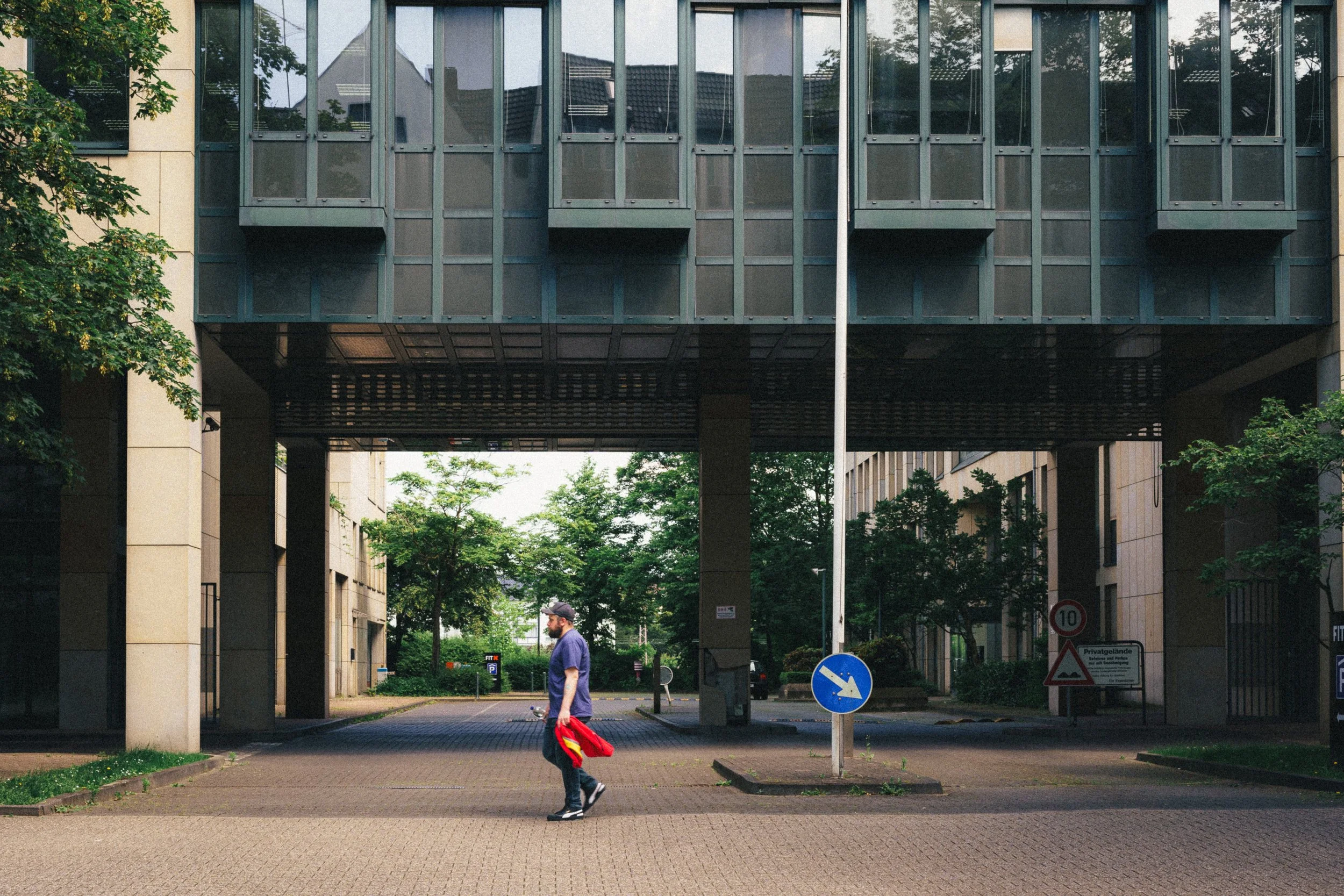 A man walking beneath modern glass and steel architecture in Düsseldorf, Germany, holding a red jacket, with urban greenery in the background, showcasing contemporary city design and everyday life.