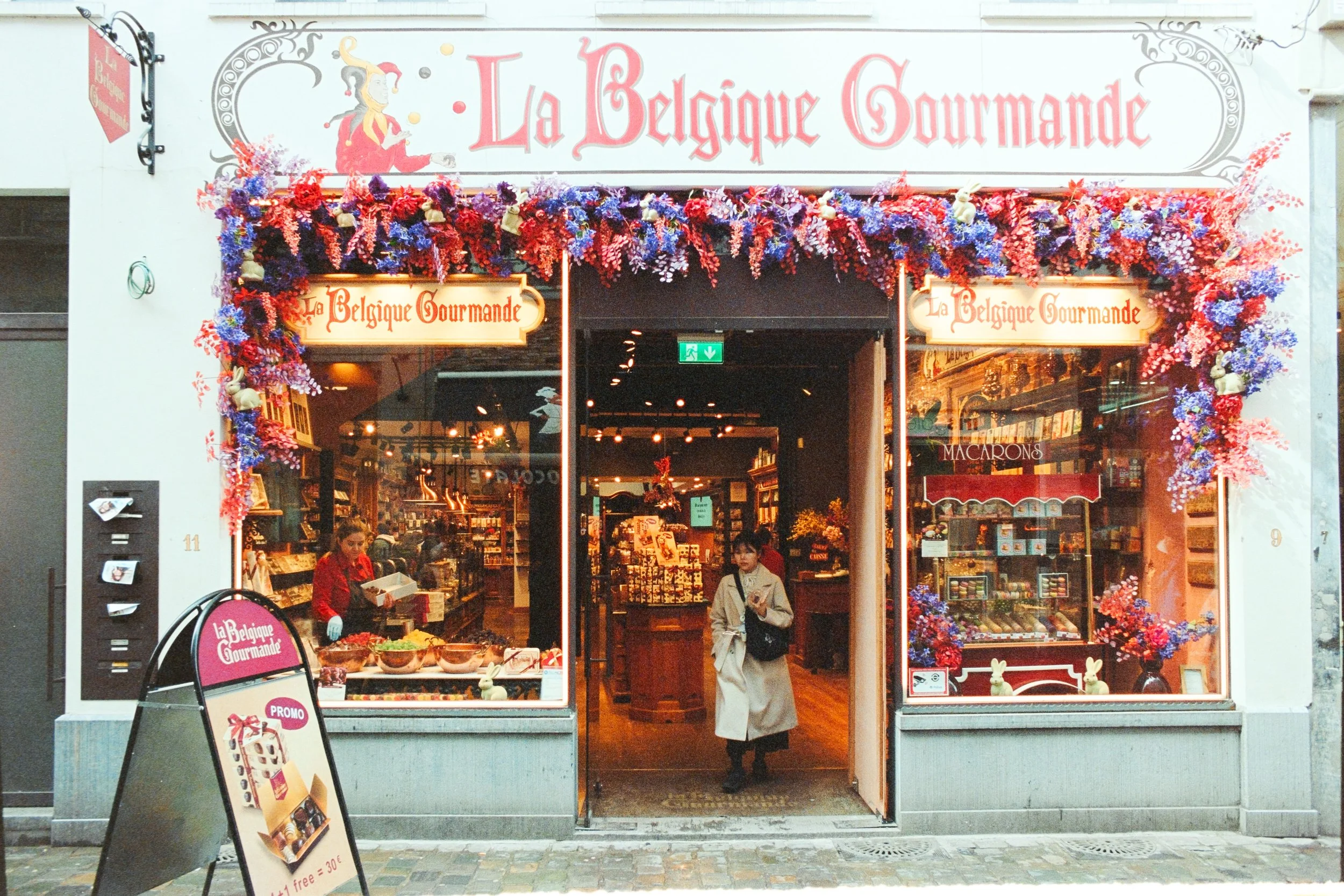 Street view of La Belgique Gourmande, a charming candy and chocolate shop in Brussels, with its entrance adorned by vibrant red and blue floral decorations. A customer in a beige trench coat exits the warmly lit shop, surrounded by displays of sweets